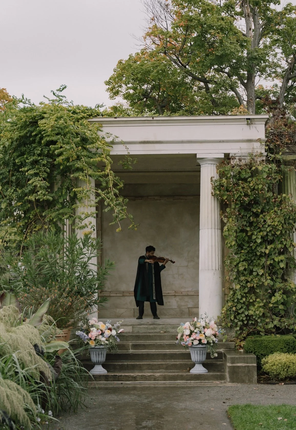 A man wearing formal black and teal clothing plays a violin on a small stage decorated with pink and white flowers, framed by tall white columns and lush greenery, with trees in the background.