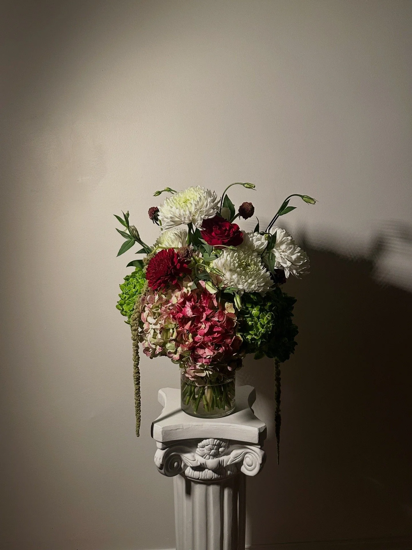 A floral arrangement with white, red, and pink flowers in a clear glass vase on a decorative white pillar, against a plain beige wall.