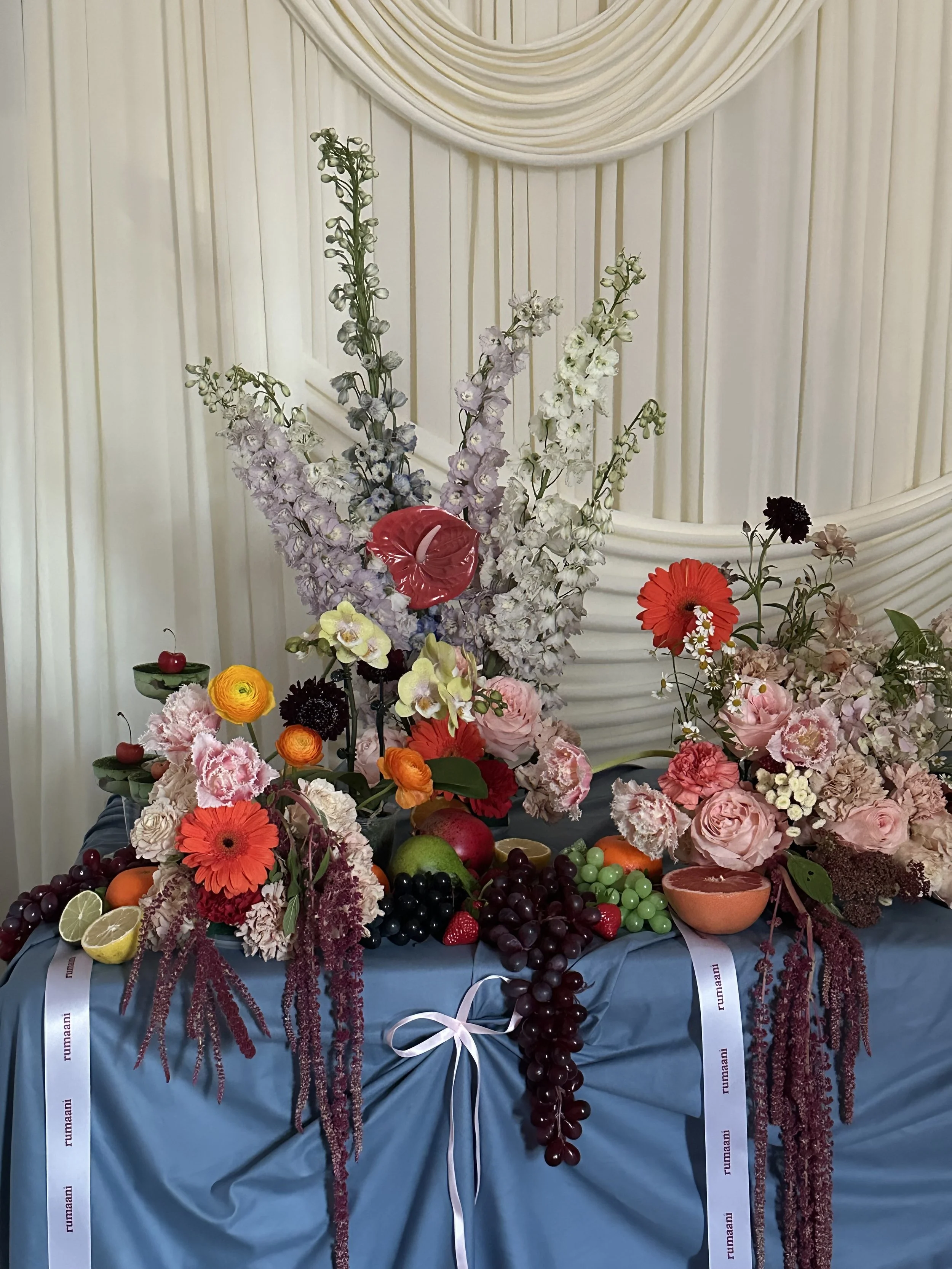 A colorful fruit and flower display on a table with a blue tablecloth and white ribbon accents, against a backdrop of cream-colored draped curtains.