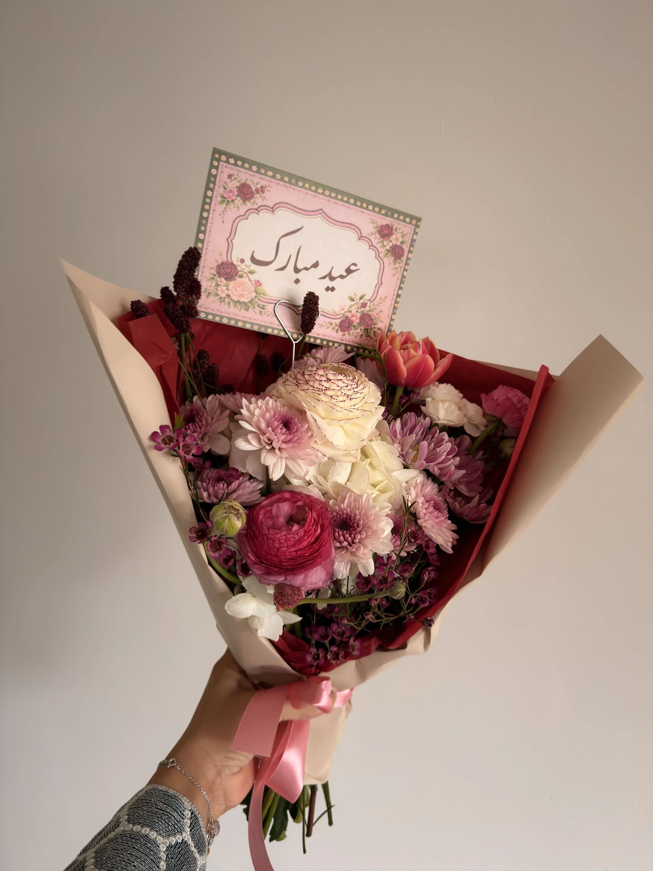 A bouquet of pink, white, and purple flowers with a pink ribbon around the stems, and a card with Persian script that says "Happy Eid."