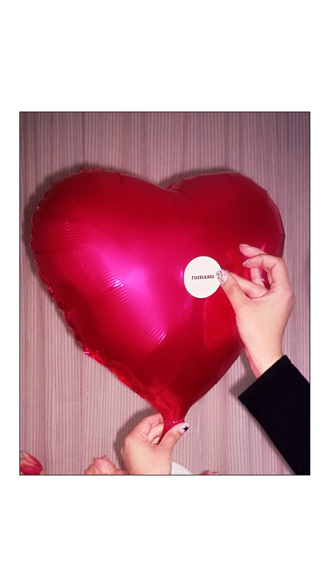 Person holding a red heart-shaped balloon with a white sticker that says 'rumaani' in front of a pink striped wall.
