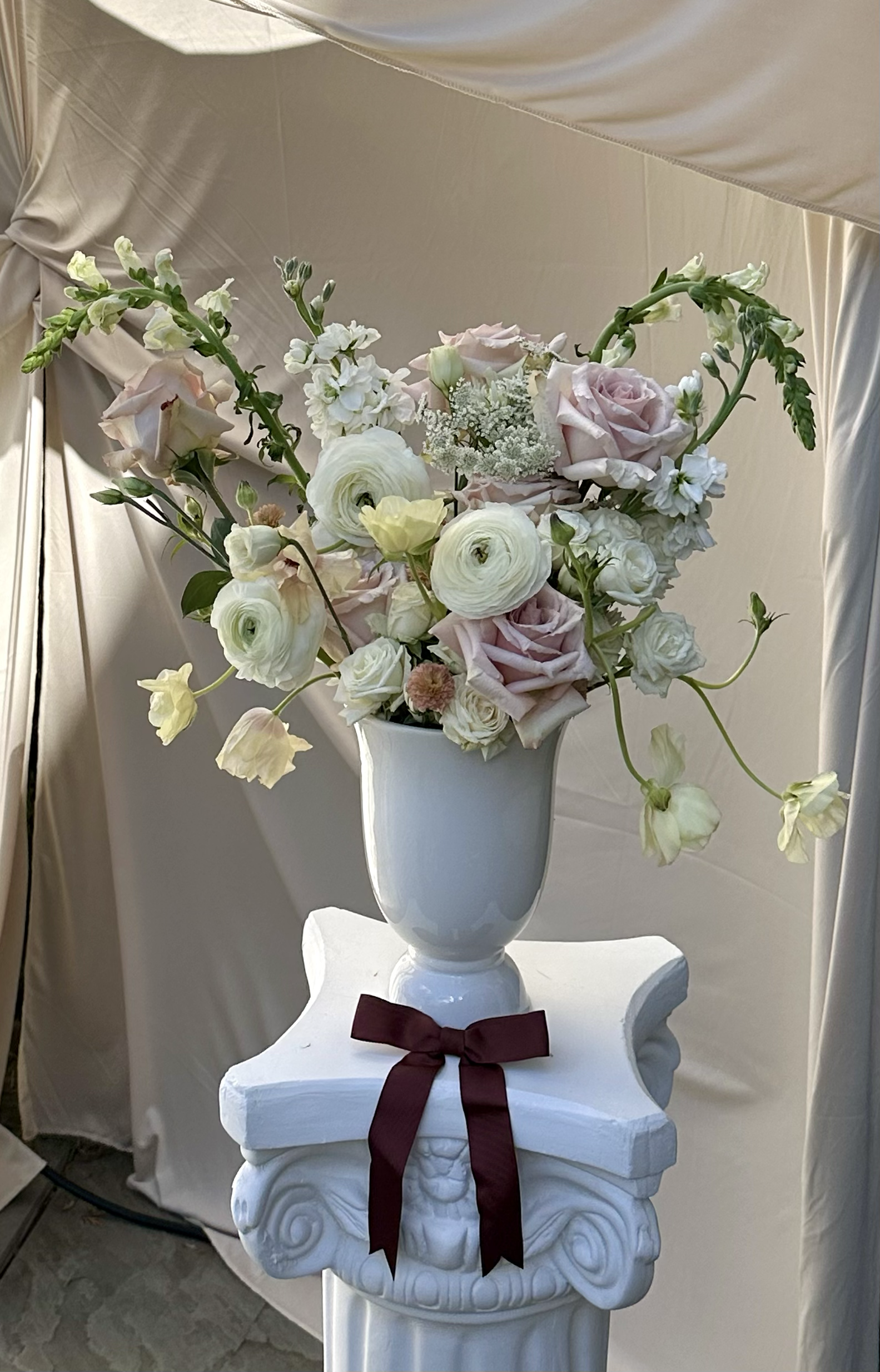 A white ceramic vase filled with light pink and white flowers, including roses and other blossoms, sits on a decorative white pedestal with a dark red ribbon tied around it. A cream-colored curtain with a draped design is in the background.