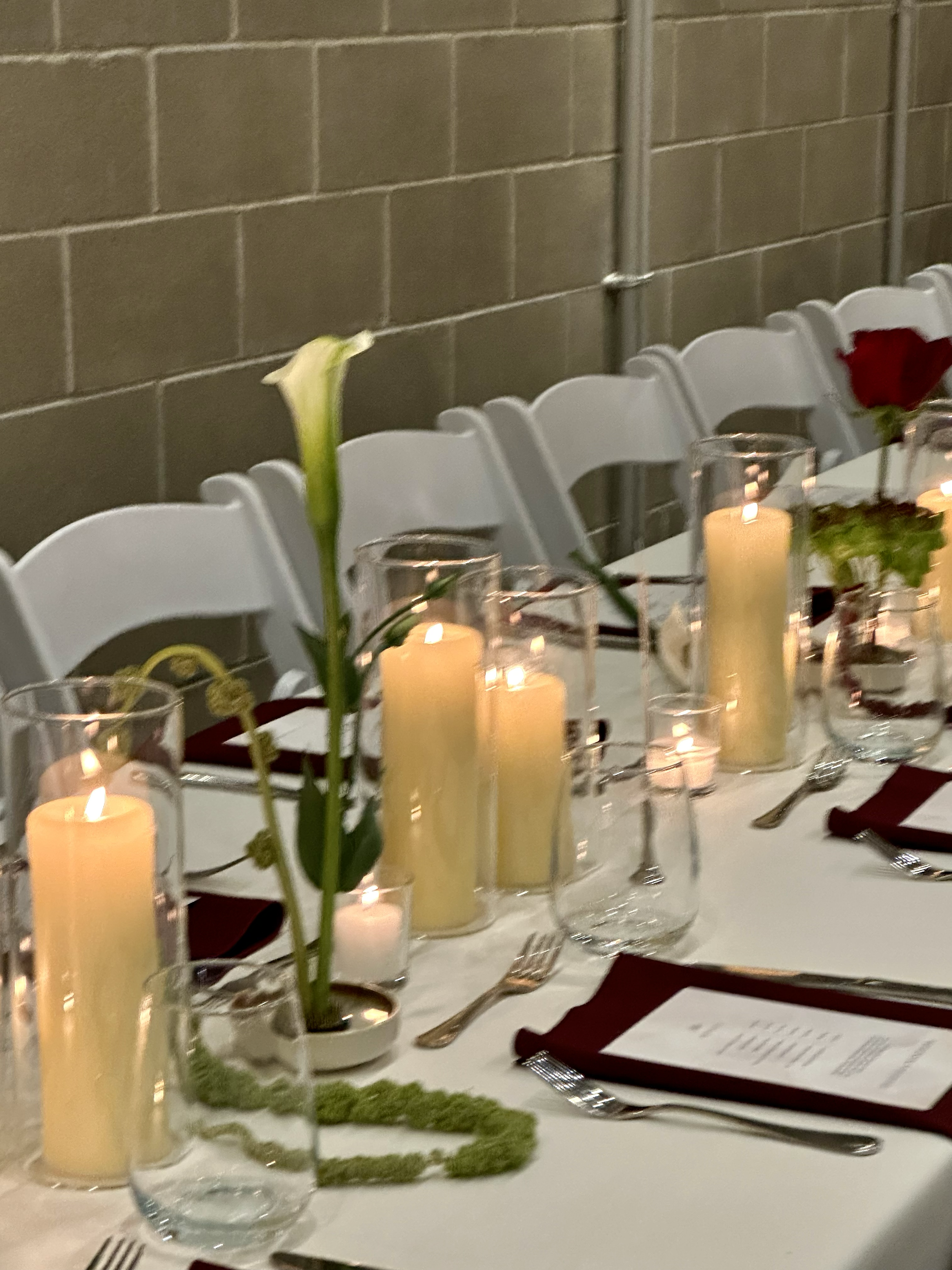 Table setting with candles, tall white calla lily in a small pot, glasses, silverware, and menus for a formal event, against a tiled wall.