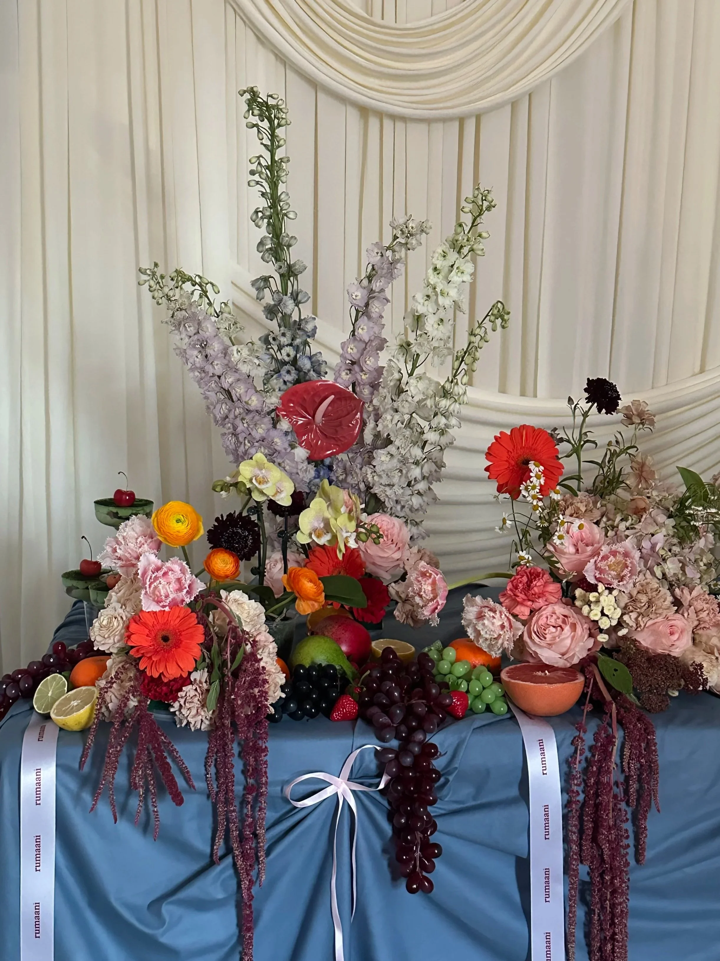 A large floral arrangement with various colorful flowers and fruits on a table, decorated with white ribbons, in a setting with a white draped backdrop.