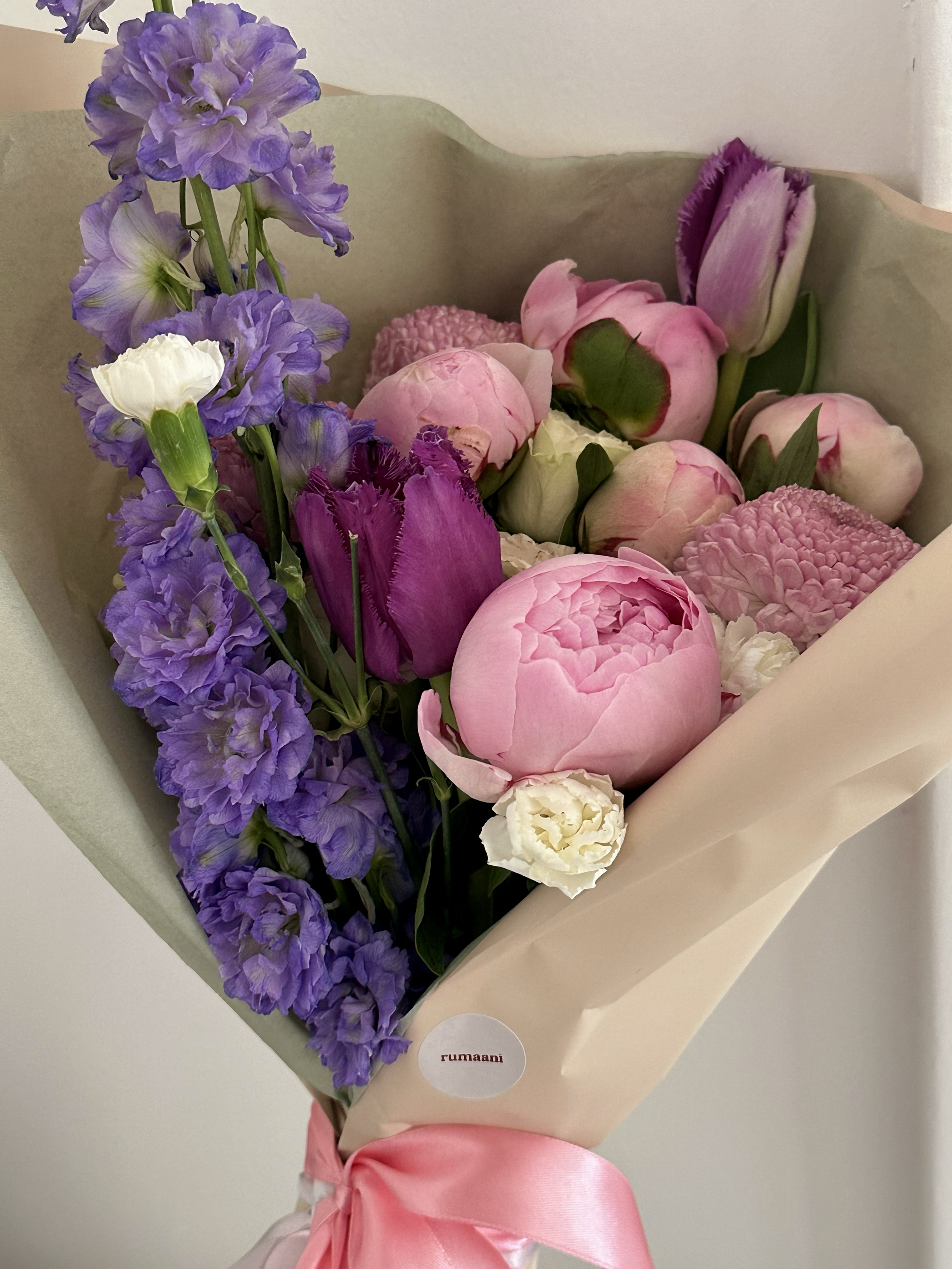 A bouquet of purple, pink, and white flowers wrapped in light paper with a pink satin ribbon and a label that reads "rumaani."