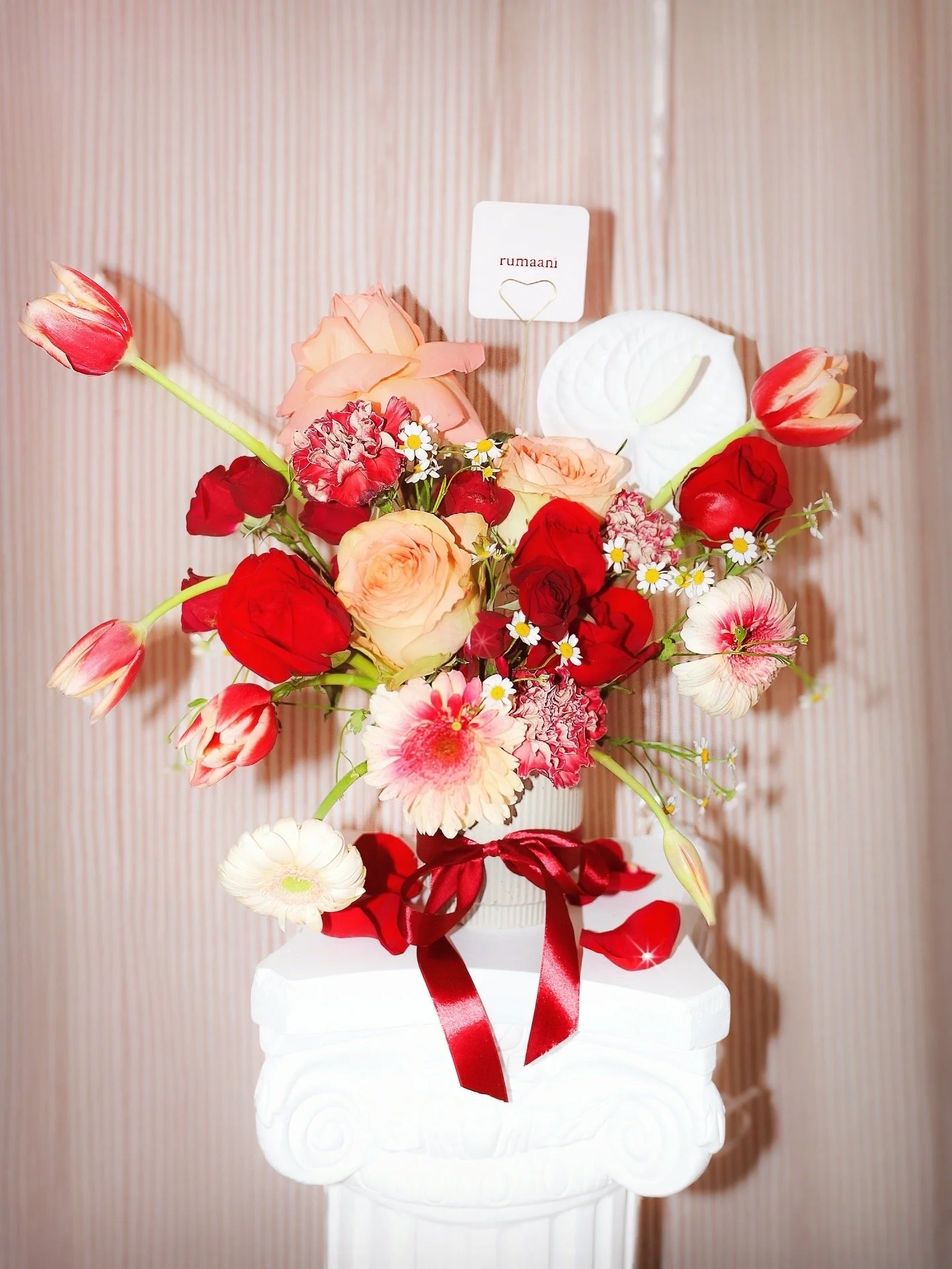 A bouquet of various red, pink, and white flowers with a small sign labeled 'rumaani' attached with a heart-shaped clip, all arranged in a white container with a red ribbon, set against a vertical cream and pink striped background.