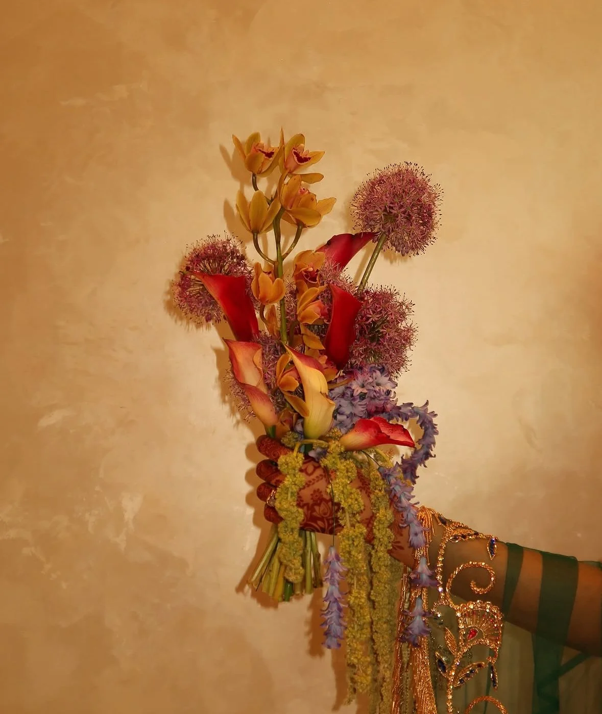 A person holding a colorful flower bouquet with various flowers including calla lilies, and other flowers in purple, yellow, and pink, with the person's arm decorated with gold jewelry and wearing a patterned green and orange outfit.