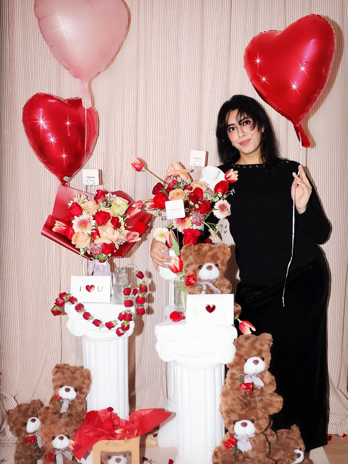 A woman standing next to Valentine's Day decorations, holding a bouquet of flowers, with heart-shaped balloons and teddy bears.