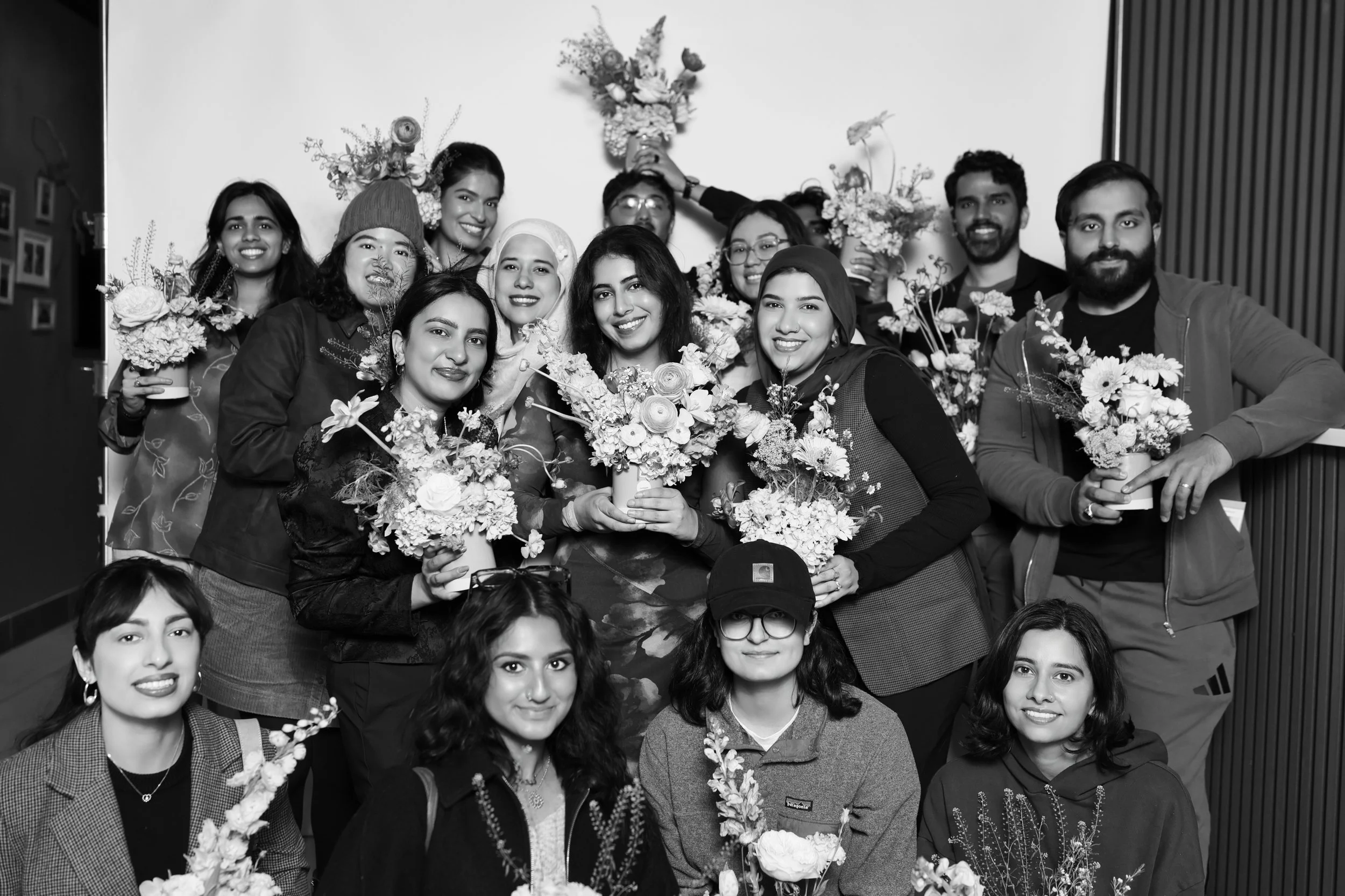 A black-and-white photo of a group of 12 diverse people smiling and holding bouquets of flowers, gathered indoors.