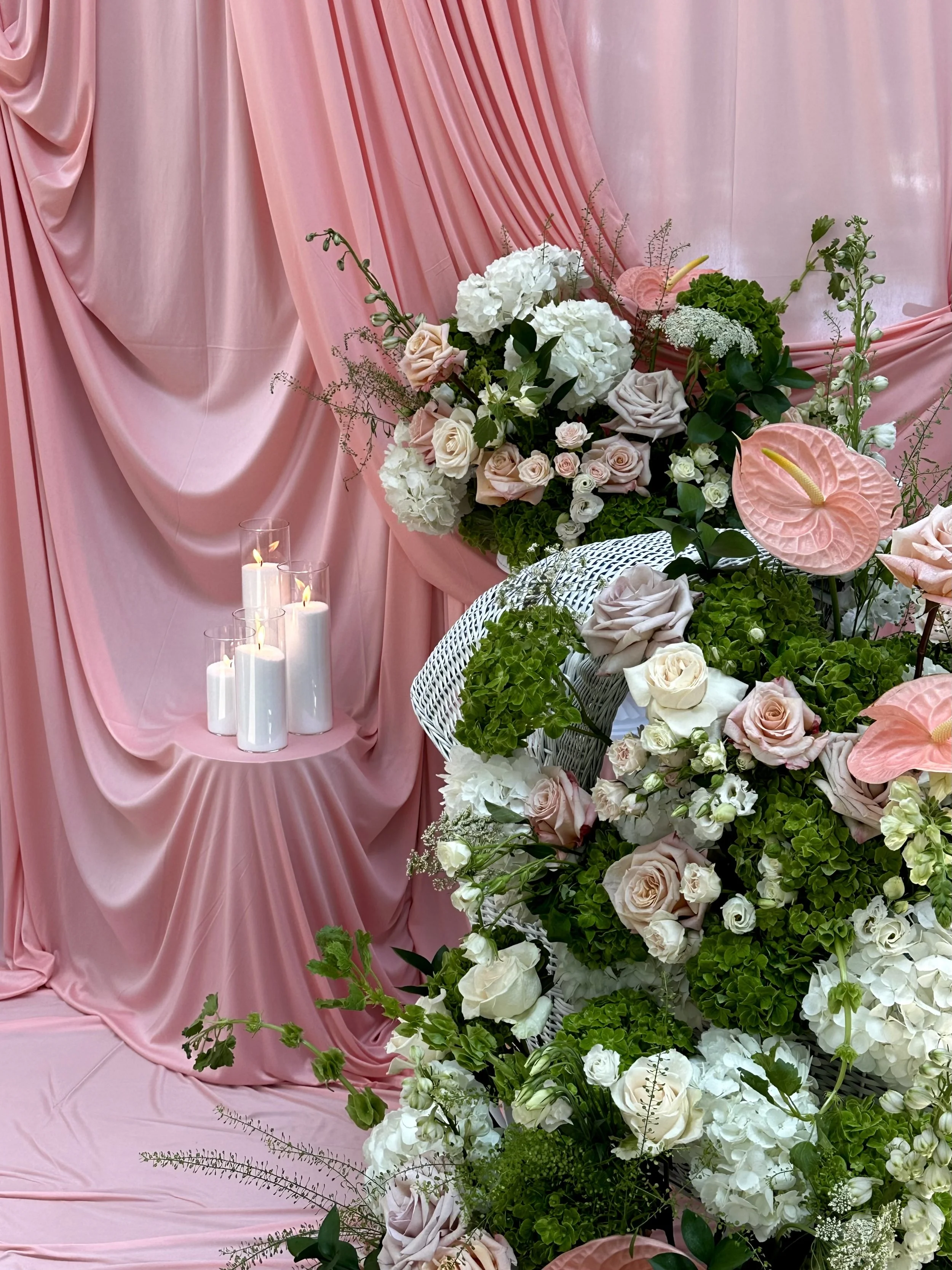 Pink draped fabric backdrop with a floral arrangement of white, blush, and green flowers, and white candles.