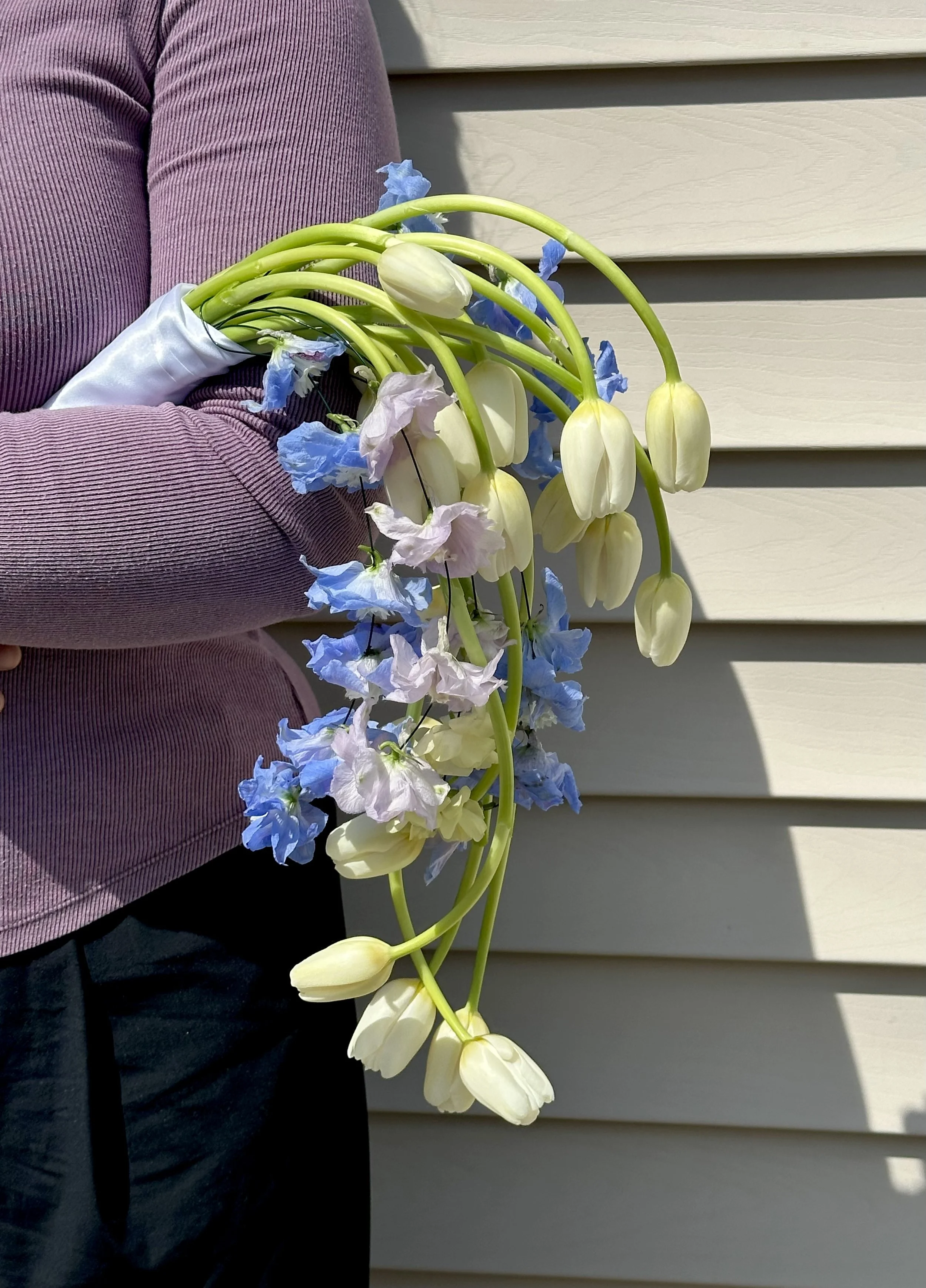 Person holding a bouquet of white and light pink flowers, standing in front of beige horizontal siding.