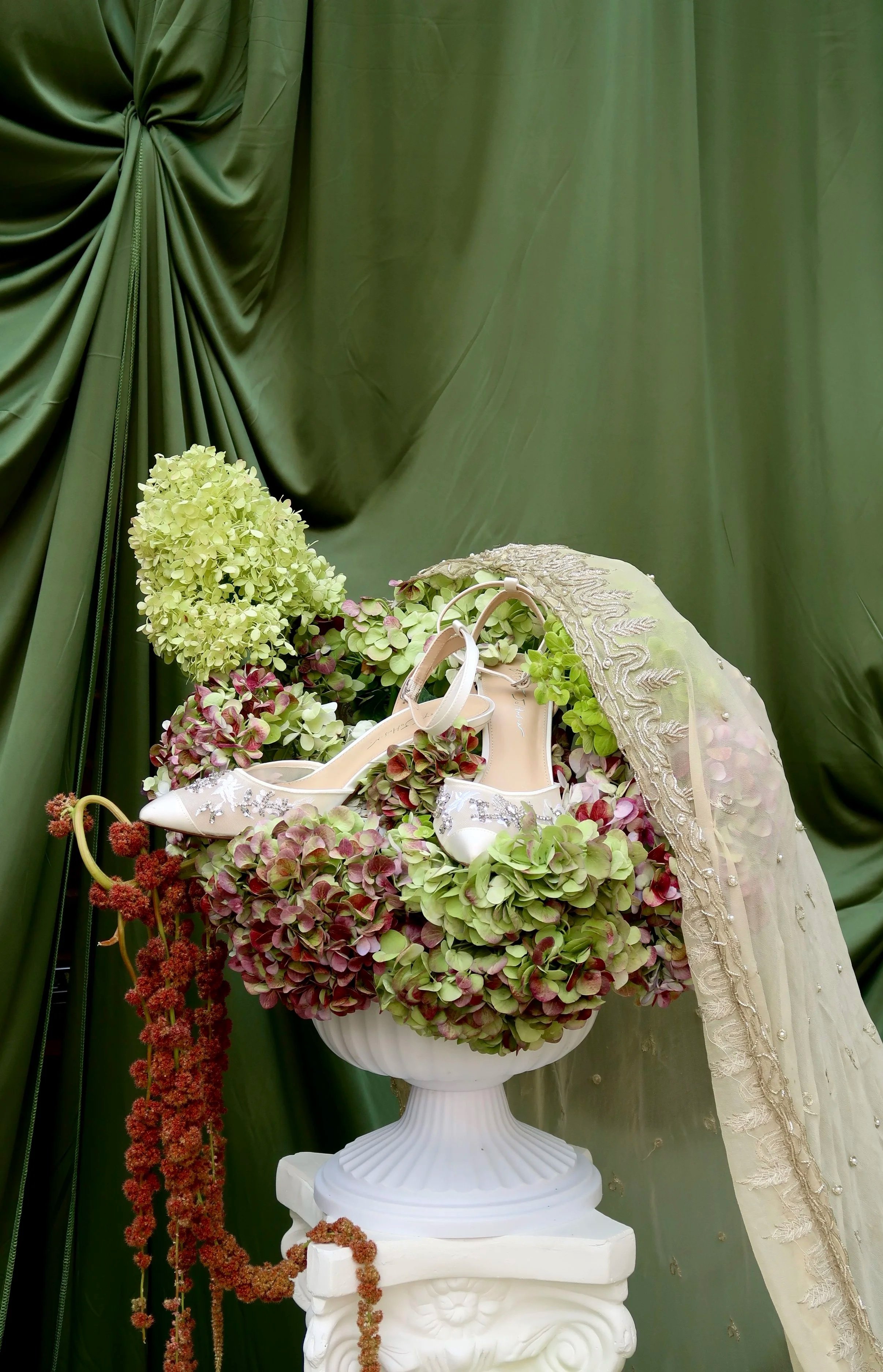 White high-heeled shoes with flower embellishments resting on pink and green hydrangea flowers in a white urn, with a lace and embroidery veil and green curtains in the background.