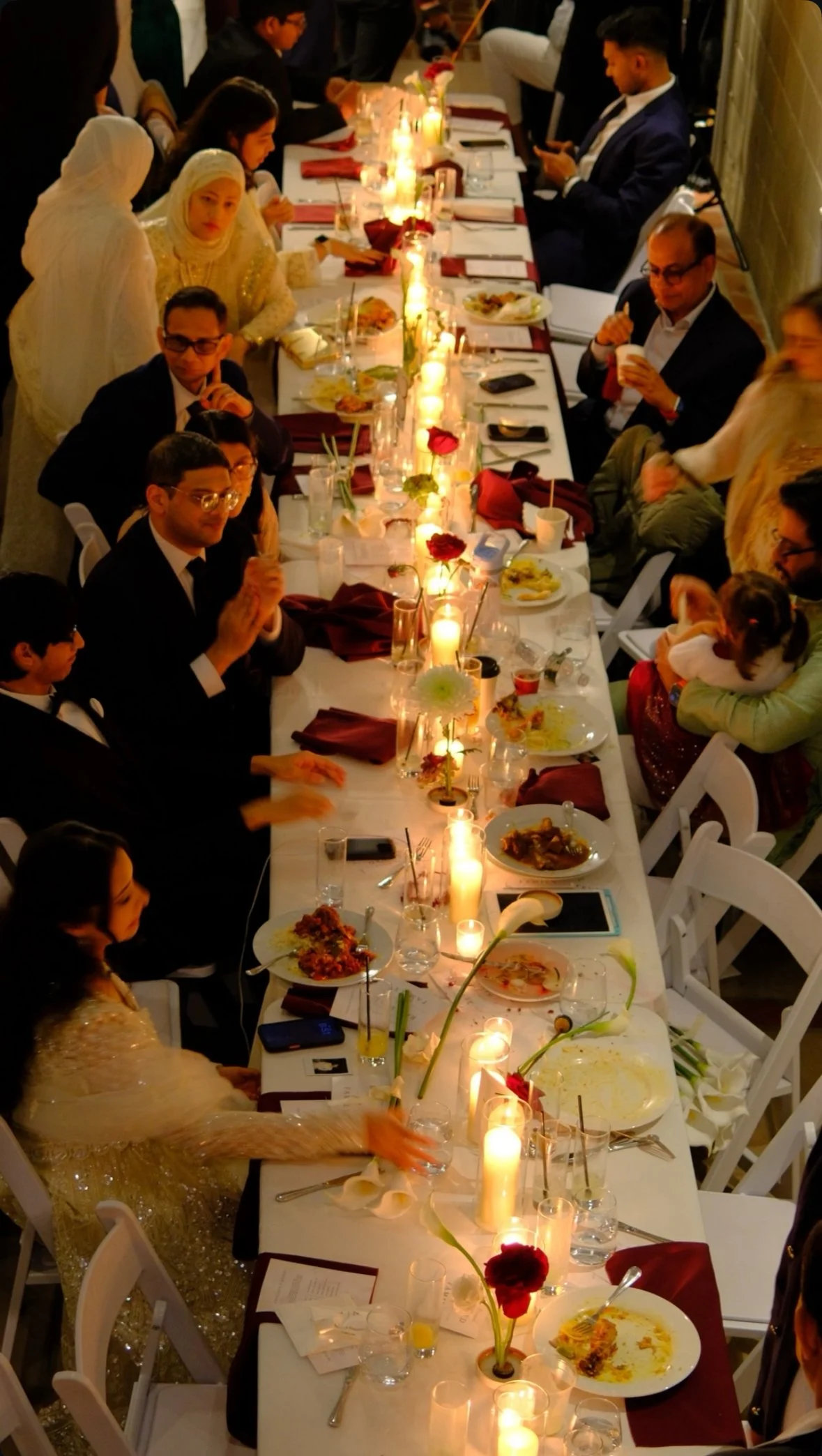 Long rectangular dinner table with guests, lit candles, flowers, and partially eaten food during a formal event or celebration.