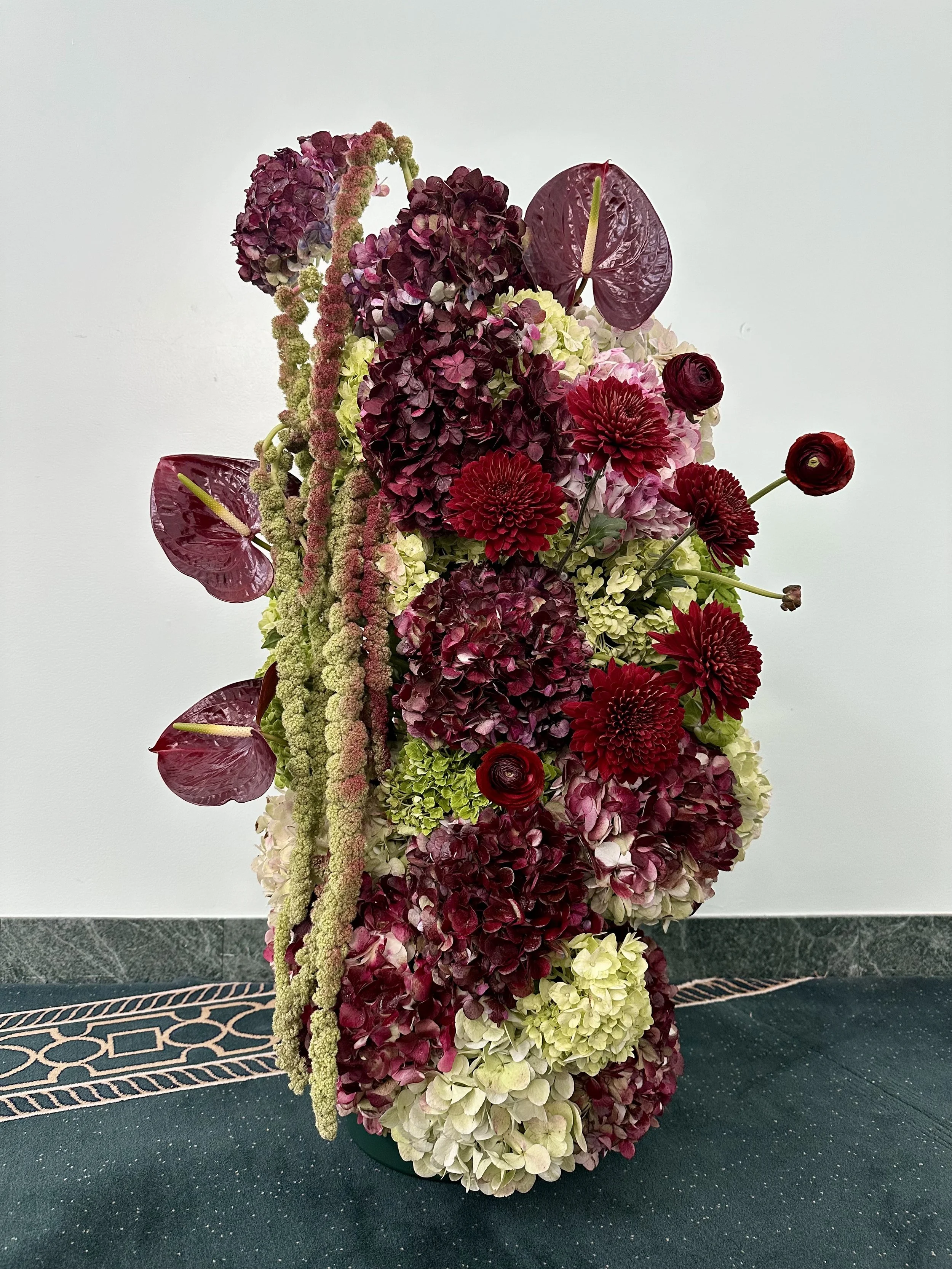 A tall floral arrangement with purple, pink, red, green, and white hydrangeas, ranunculus, red dahlias, and anthuriums against a plain white wall.