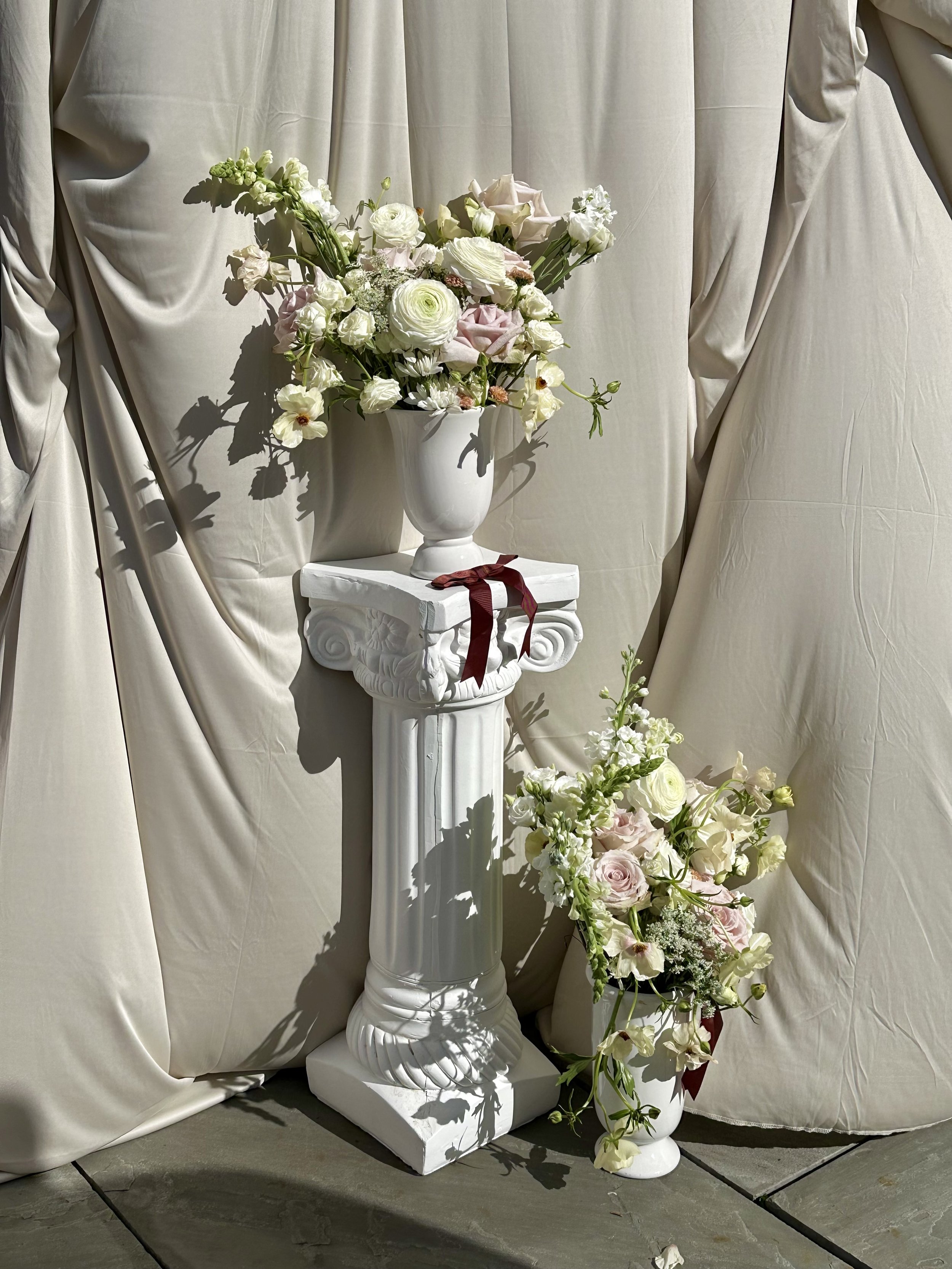 A white floral arrangement in a white vase on a white pedestal, set against a cream-colored draped fabric background.