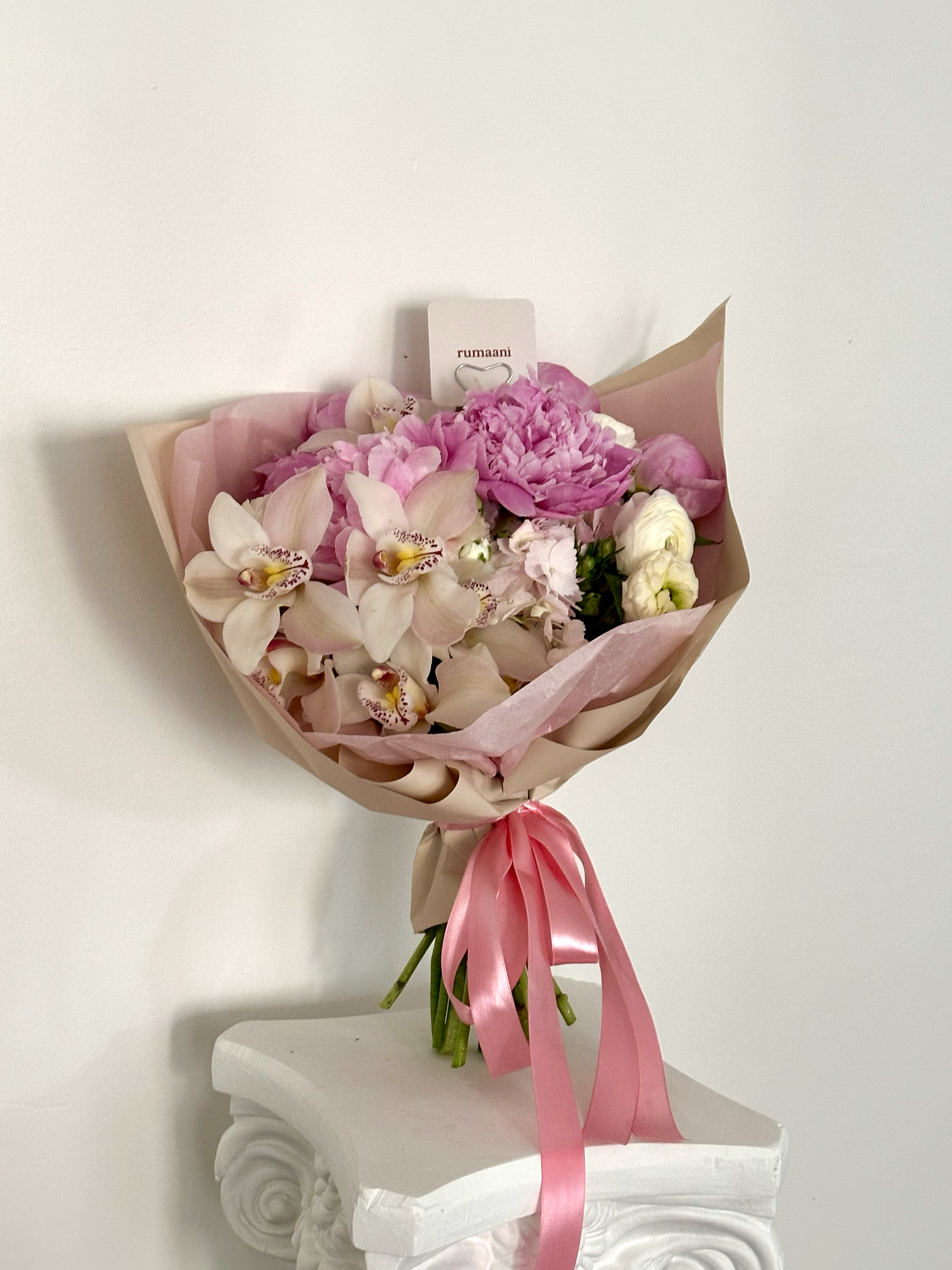 A bouquet of pink and white flowers with a pink ribbon, resting on a white decorative pedestal against a plain white background.