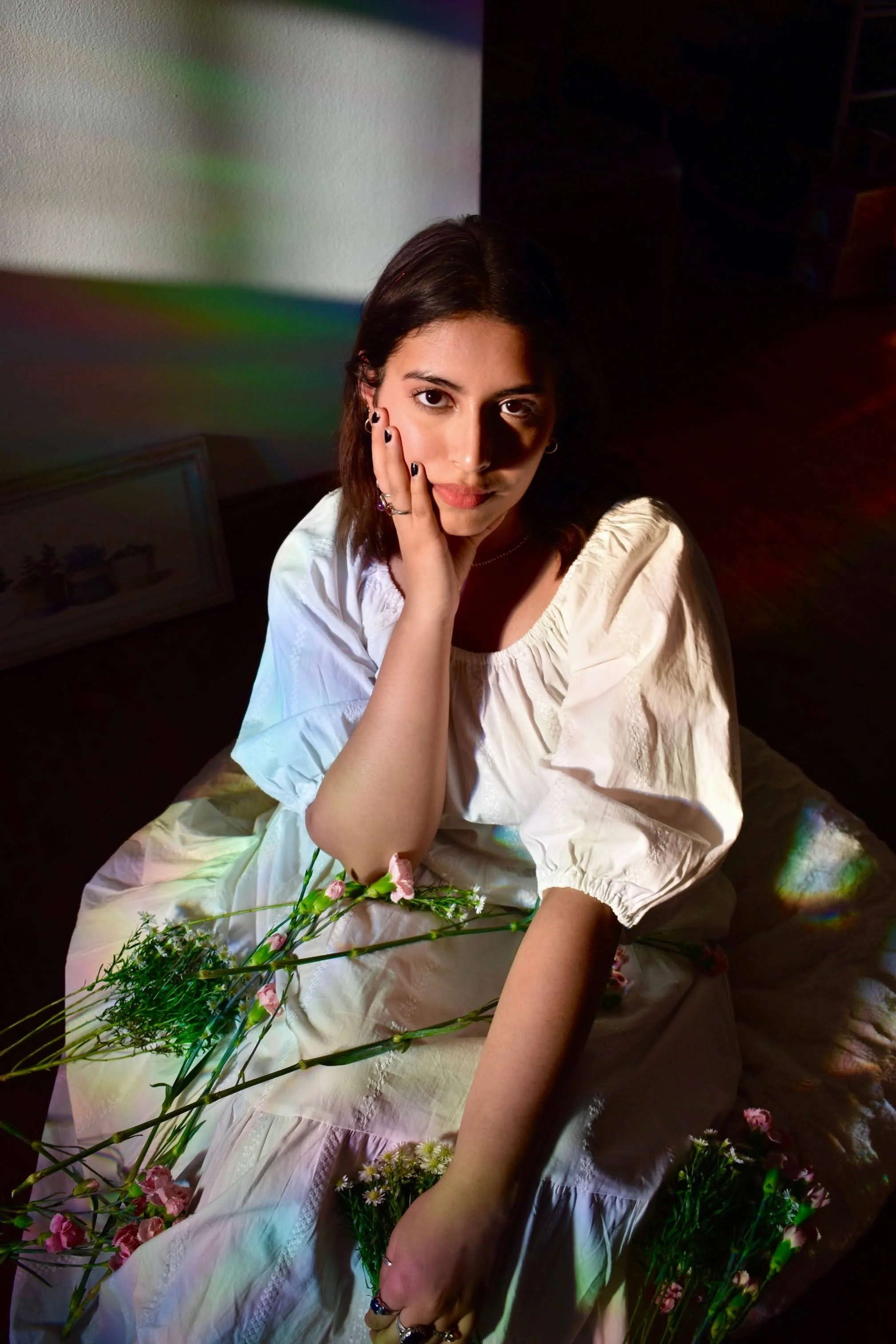 A young woman with dark hair and earrings, wearing a cream-colored dress, sitting amongst pink and white flowers, holding her face with one hand, and looking directly at the camera. The background is partially dark with some light and rainbow-like reflections.