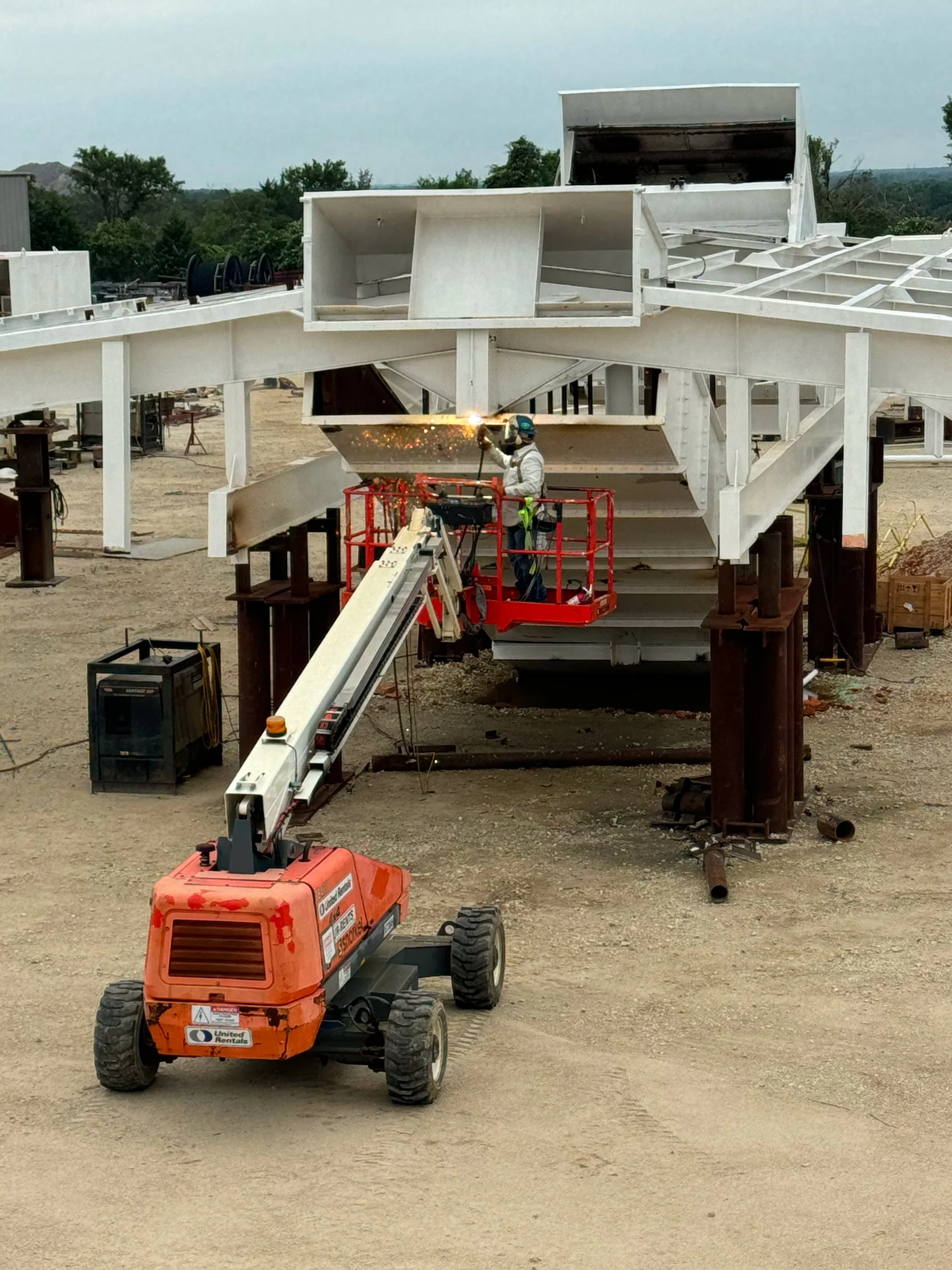 A construction worker welding a large metal structure at a construction site, using a cherry picker lift for elevation. The site has various construction equipment and materials on a dirt ground, with trees and a cloudy sky in the background.
