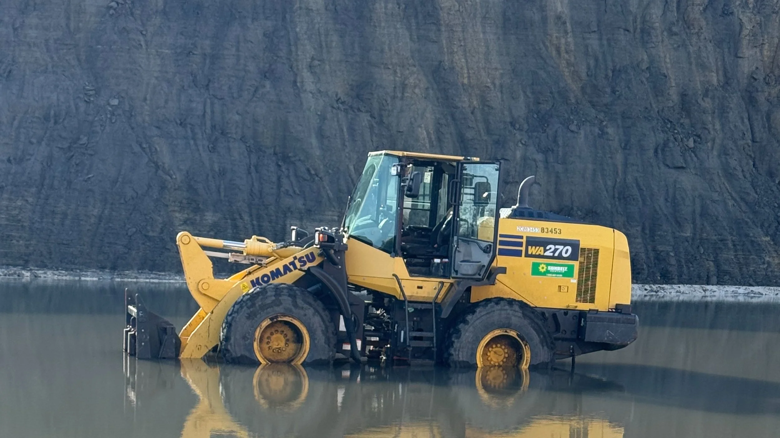 Yellow Komatsu WA270 bulldozer operating in a muddy area with a large dark cliff in the background.