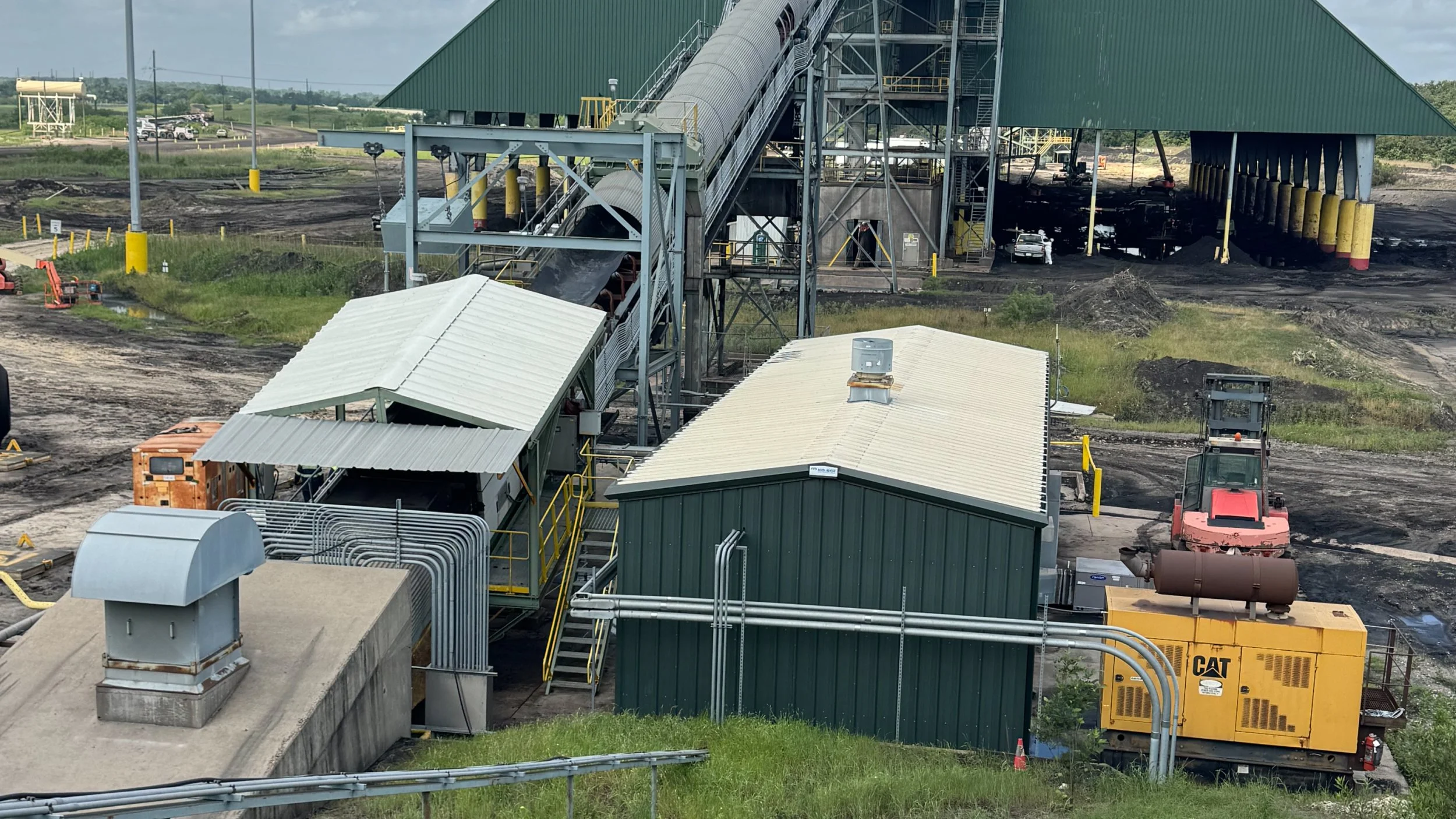 Industrial site with conveyor belts, small buildings, construction equipment, and dirt pathways, likely for processing or manufacturing.