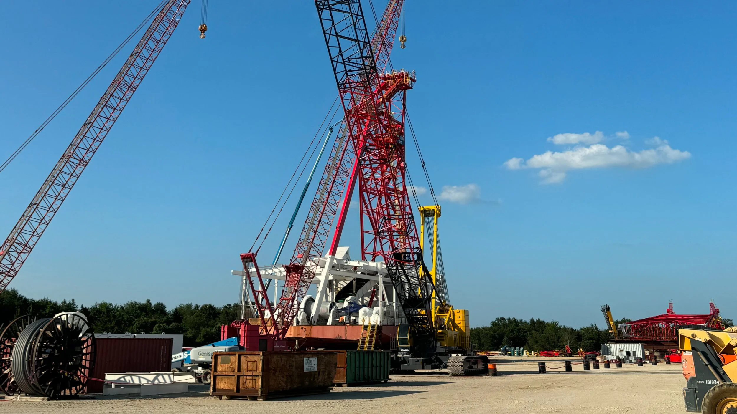 Construction equipment yard with large cranes, machinery, and supplies under a clear blue sky.