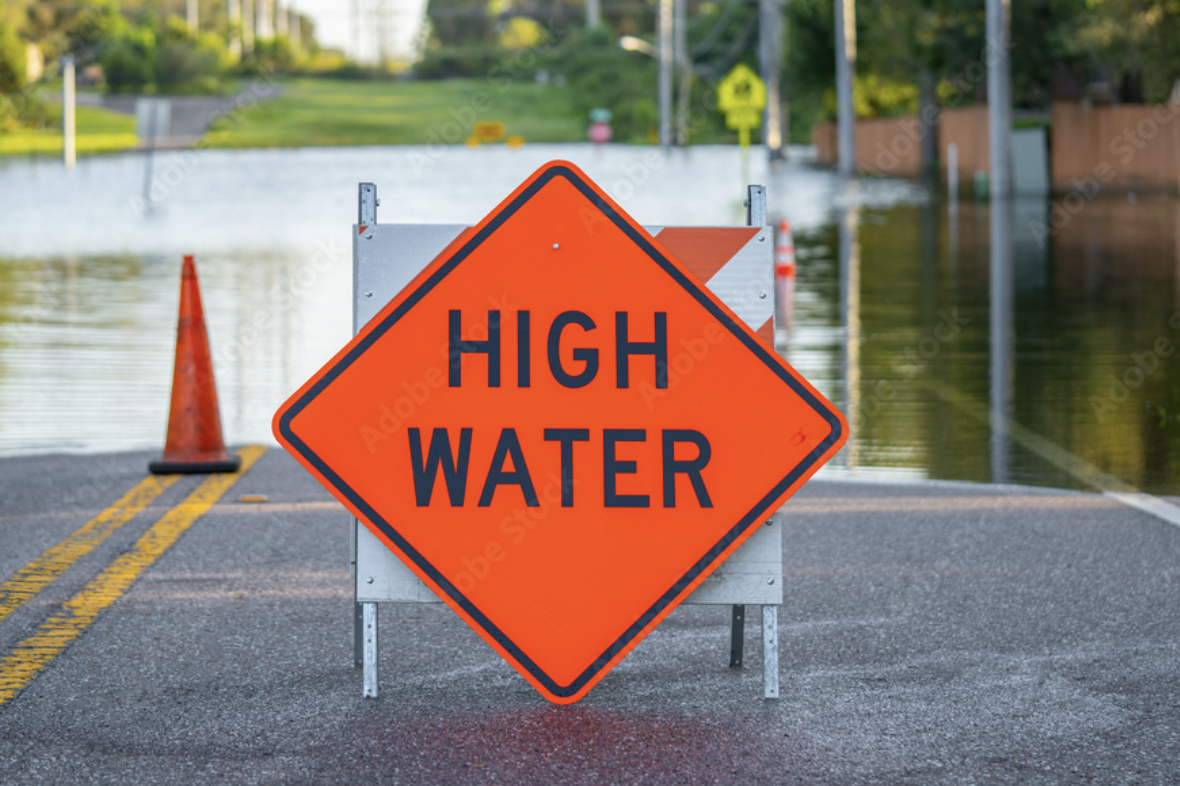 A bright orange road sign with black text that reads "High Water," indicating flooding or high water levels on the road, with cones and barriers blocking the roadway.