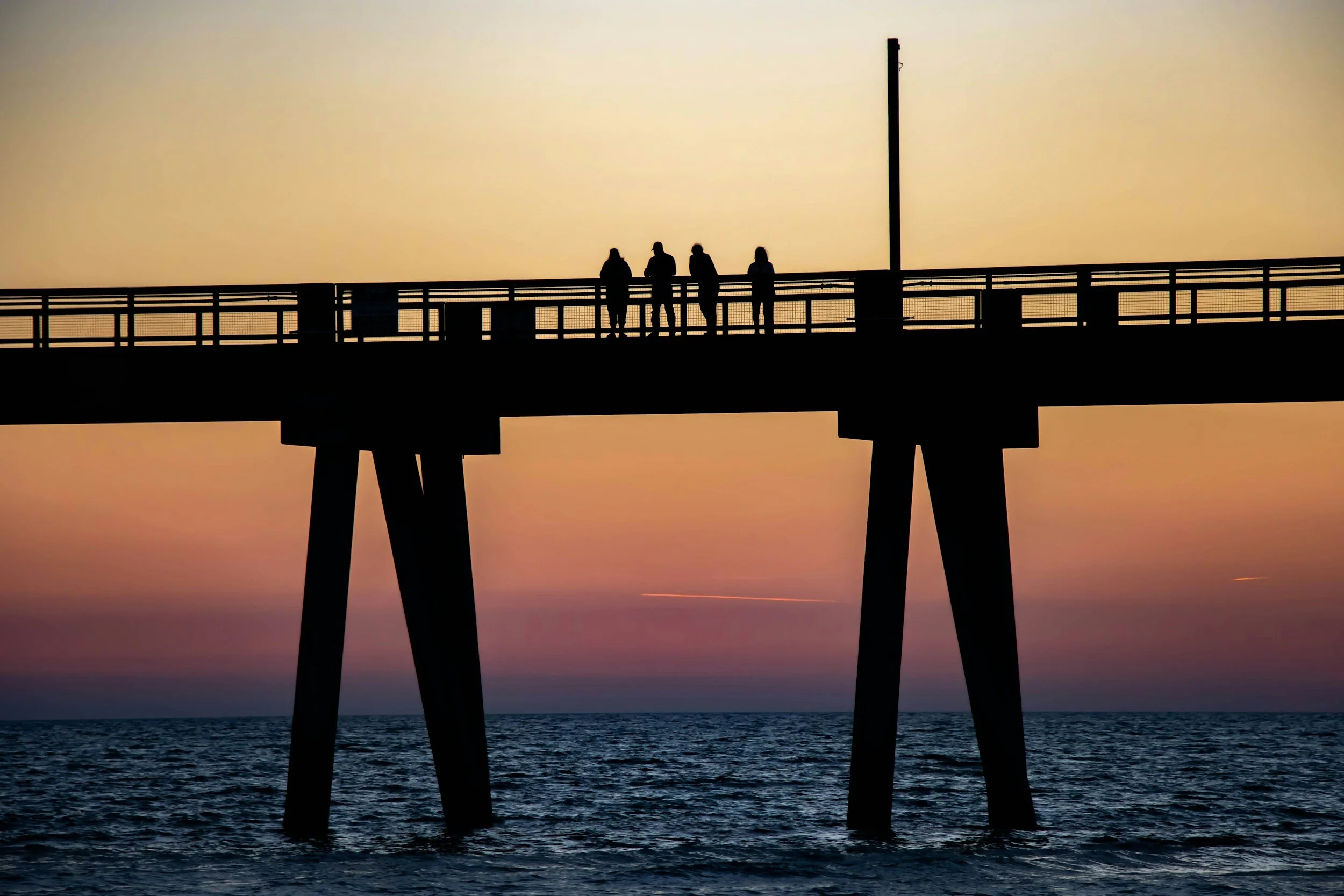 Silhouettes of four people standing on a pier in Florida at sunset over the ocean, with a tall pole on the right