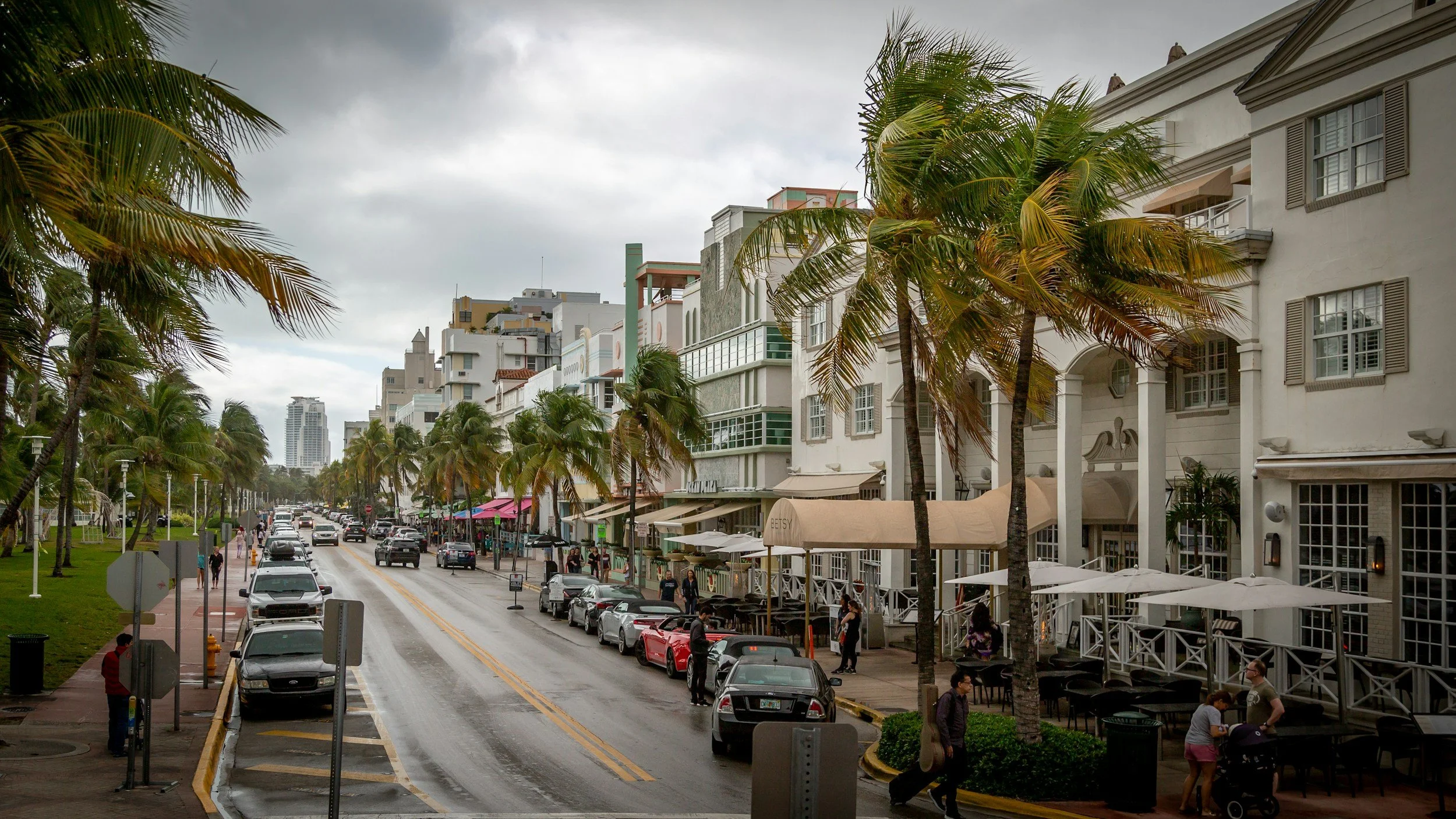 A city street in Florida lined with palm trees, with modern multi-story buildings on one side and parked cars along the curb. There are people walking on the sidewalk and outdoor seating with umbrellas for restaurants or cafes. Overcast sky overhead.