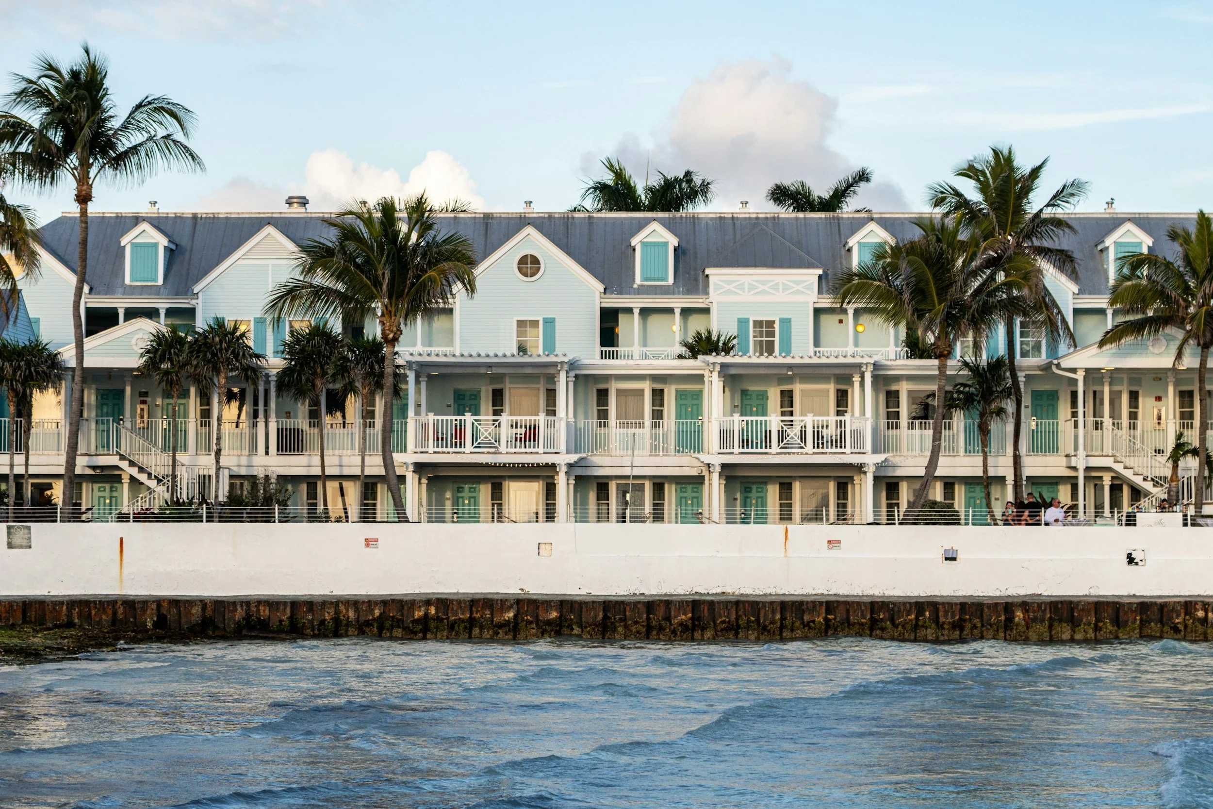 A multi-story building with teal doors and shutters, surrounded by palm trees, situated along a waterfront with a retaining wall and water in the foreground.