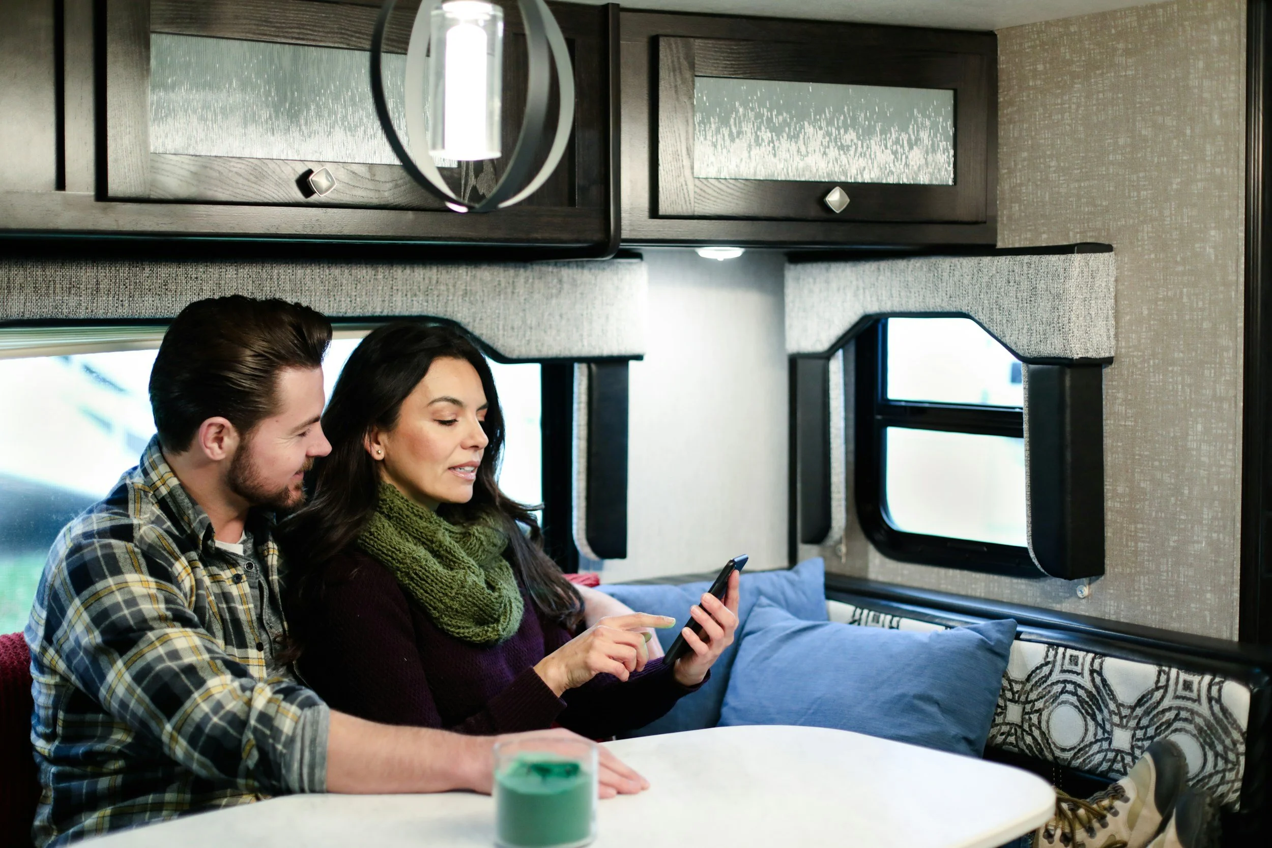 A man and woman sitting close together on a couch inside an RV, with the woman holding a smartphone and the man looking at it, while the woman is also looking at her phone. There are two windows behind them and overhead storage cabinets. The woman is wearing a green knitted scarf and a dark sweater, and the man is wearing a plaid shirt.