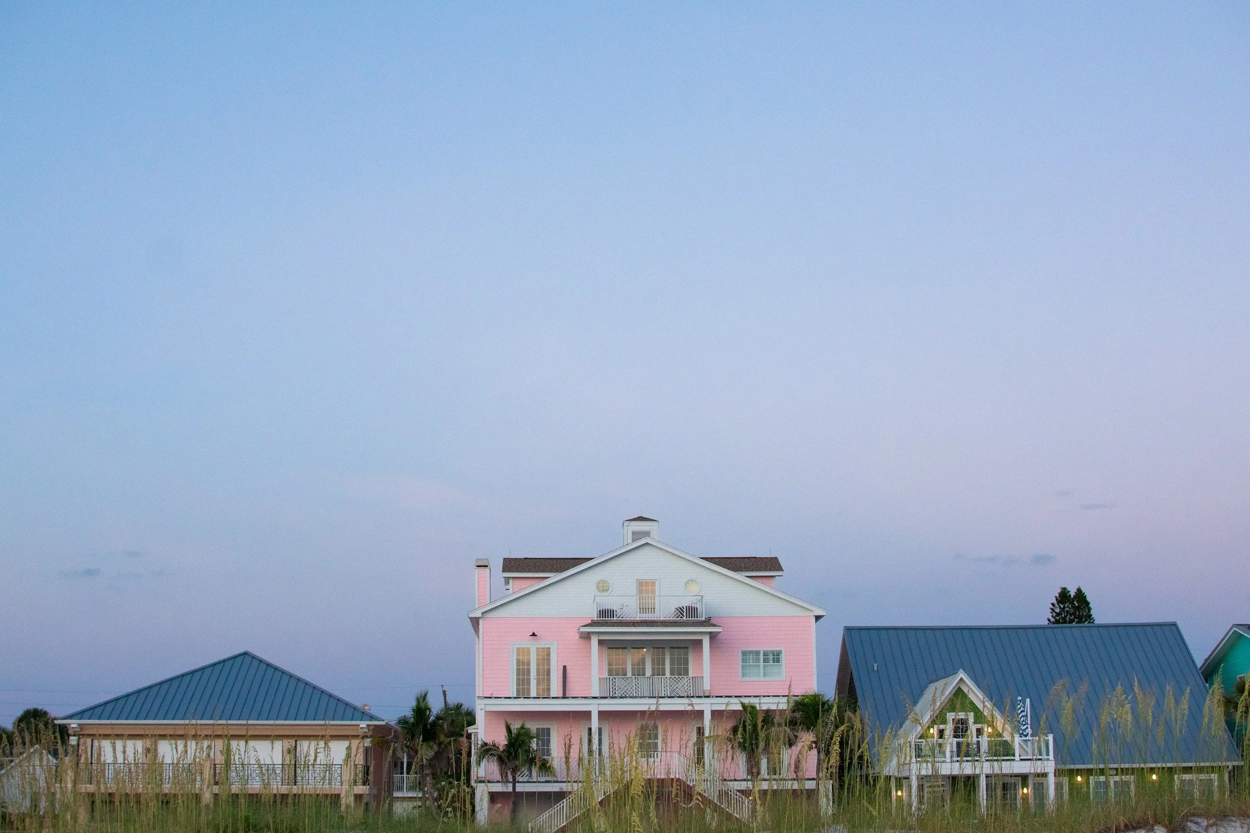Large pink house with white trim, balconies, and multiple windows, surrounded by grass and other houses with blue roofs under a mostly cloudy sky.