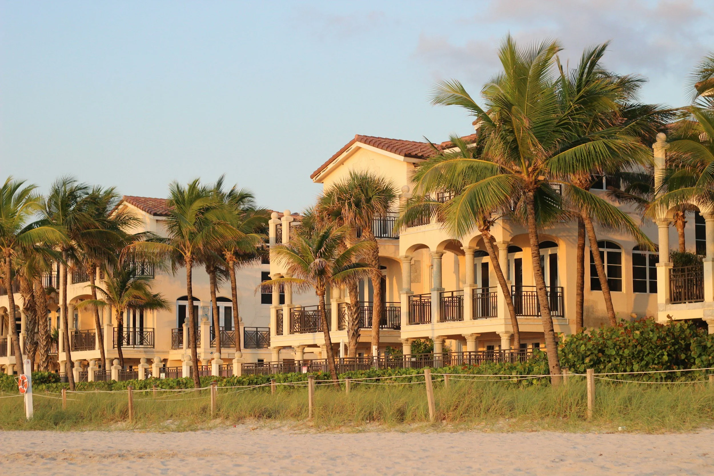 A beachside view with a grassy area, a row of palm trees, and a large white Mediterranean-style building with tiled roofs and arched windows.