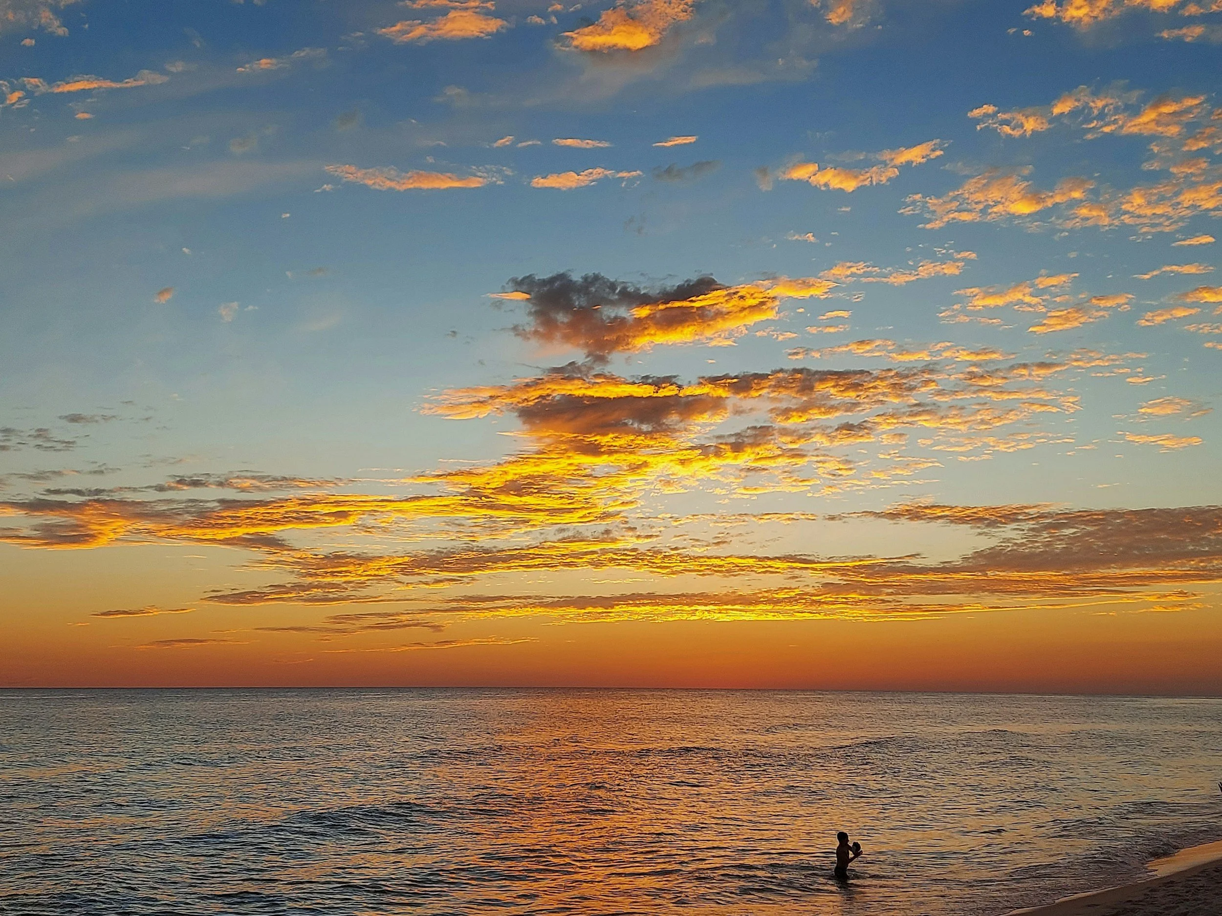 A sunset over the ocean with orange, pink, and blue clouds in the sky. Silhouetted person standing at the water's edge