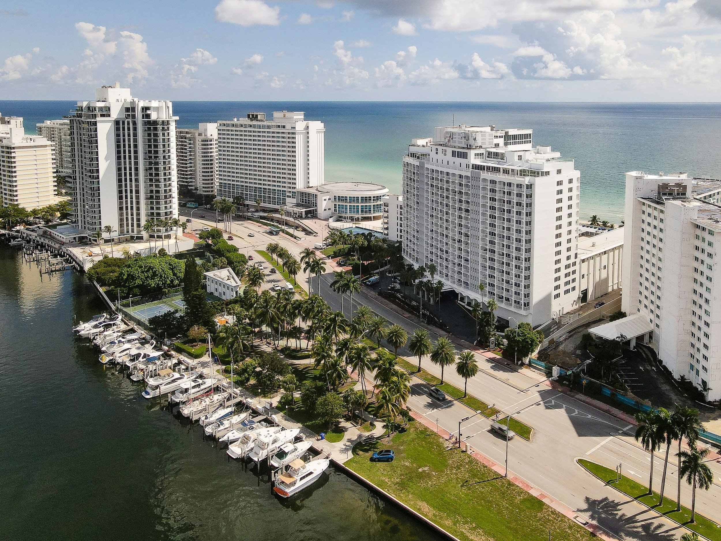 Aerial view of a coastal area with high-rise hotels and buildings along the shoreline, a marina with boats docked, and a waterway. The ocean extends into the horizon beneath a partly cloudy sky.