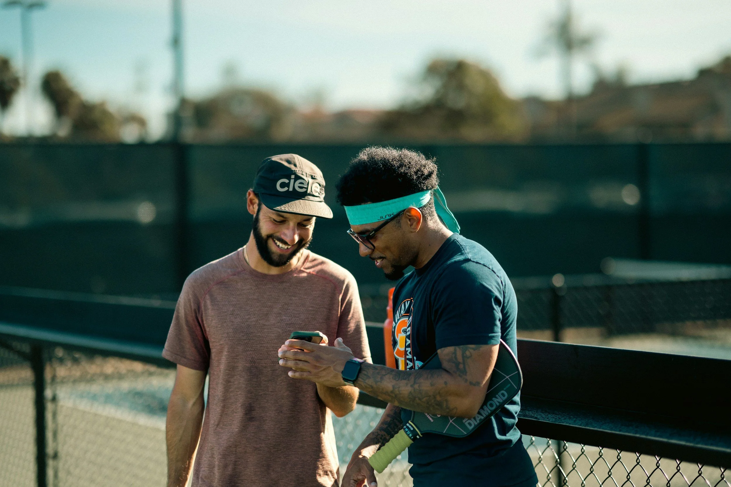 Two young men, one in a cap and the other with a headband, are standing outside on a tennis court, looking at a phone and smiling.