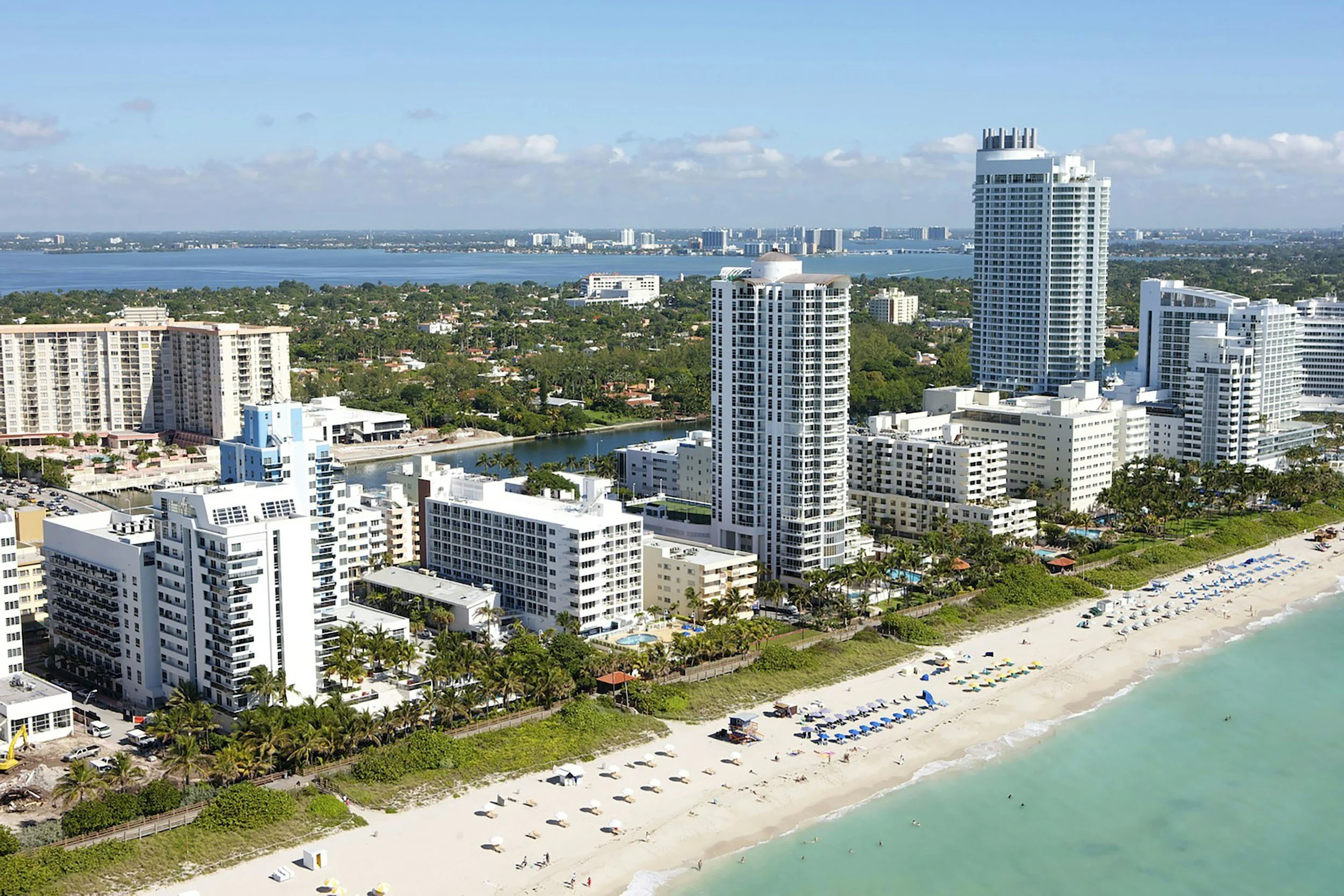 Aerial view of a coastal city with high-rise buildings near the beach, a body of water in the background, and a sandy shoreline with umbrellas and beach chairs.