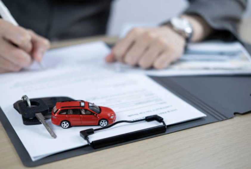 A person's hands working at a desk with car keys, a toy red car connected to a black cord, and paperwork.