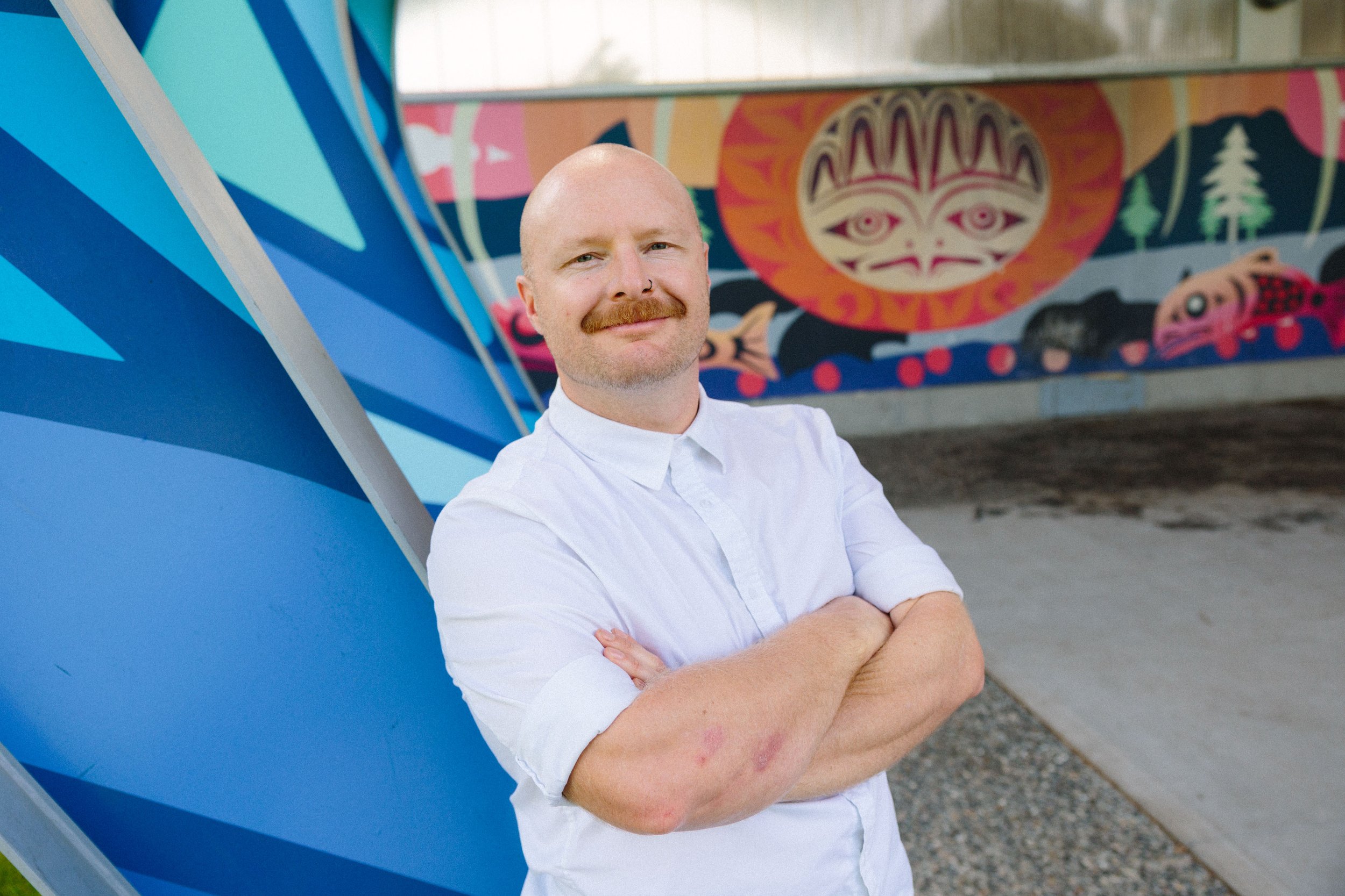 A photograph of Sean Easton - a man in his thirties with a bald head and moustache - standing in front of the community mural in O'Siyam Pavilion in Squamish.
