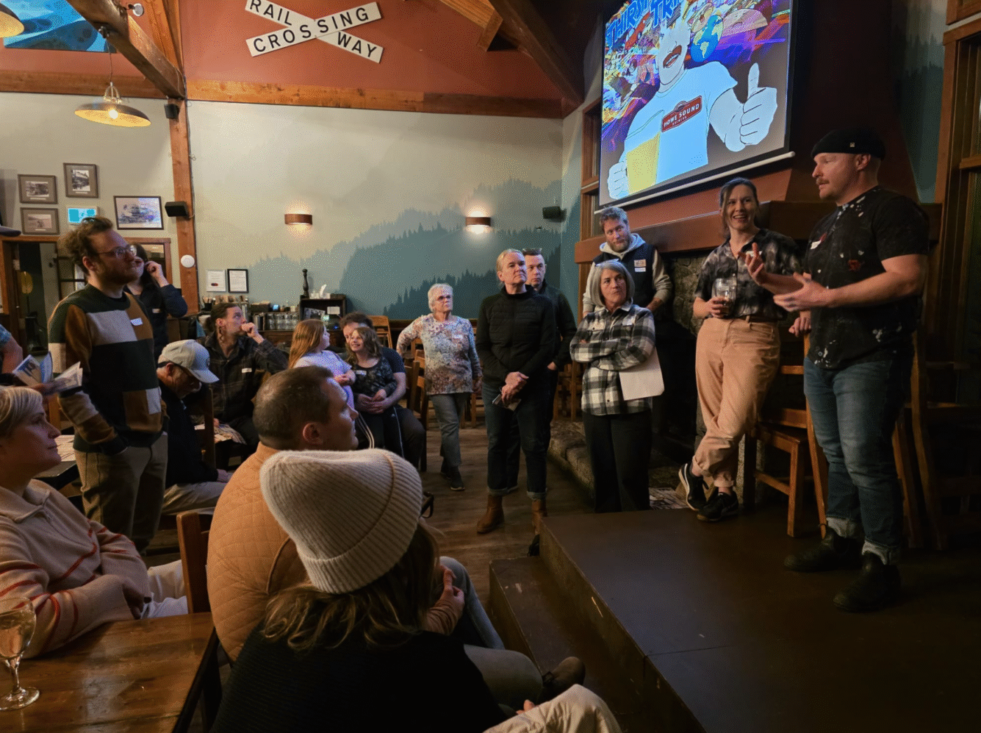 A group of people are attending a presentation or event in a cozy, rustic room with wooden beams. Several individuals are standing near a small stage, and one speaker, Sean Easton is talking. The audience is seated and standing, listening attentively