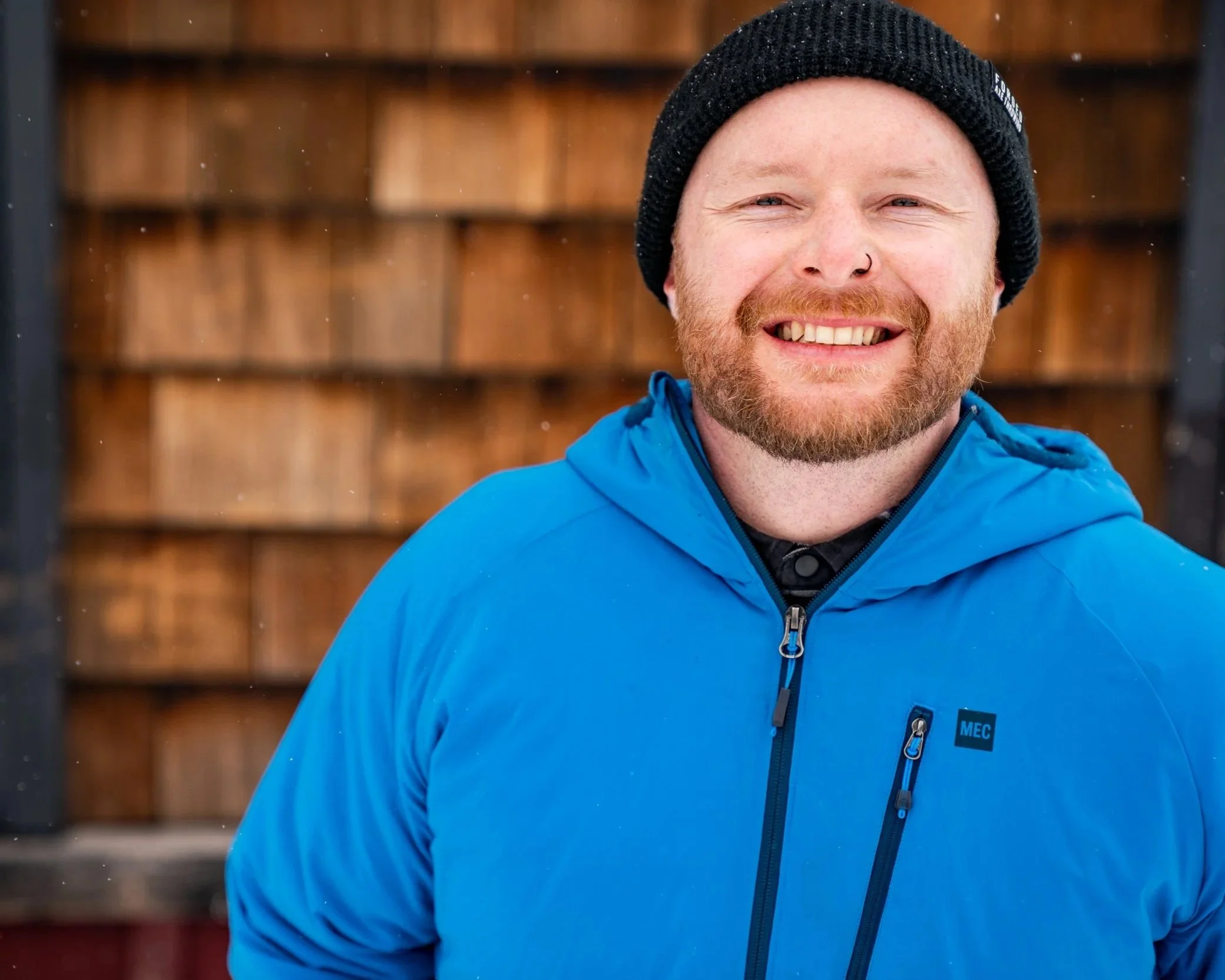 Close-up Sean, a smiling man in his 30s with a beard. He's wearing a black knit beanie and a blue jacket, with a wooden background.
