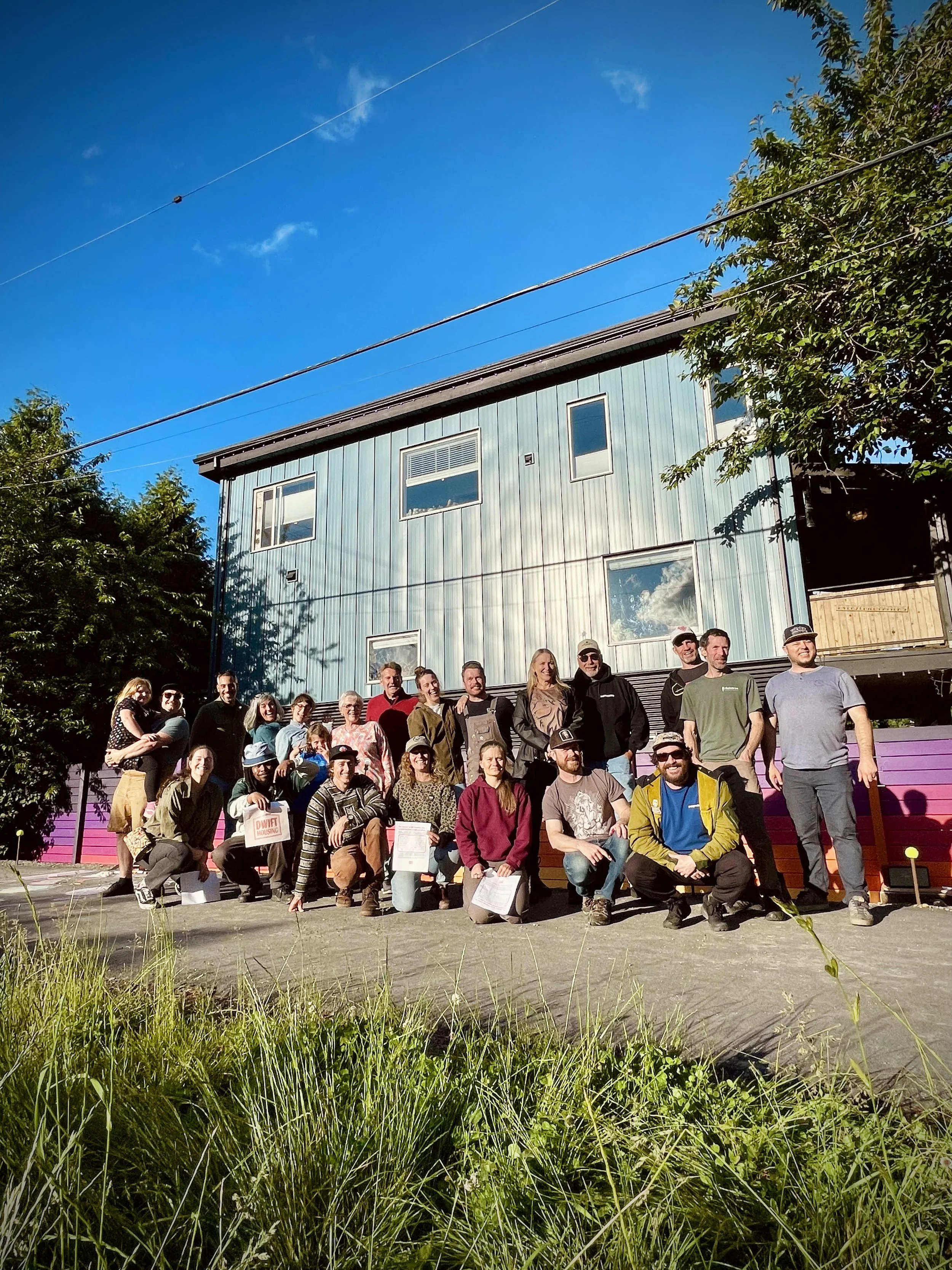 Group of people standing and sitting outside in front of a blue house with a brightly painted fence and trees, and a clear blue sky.