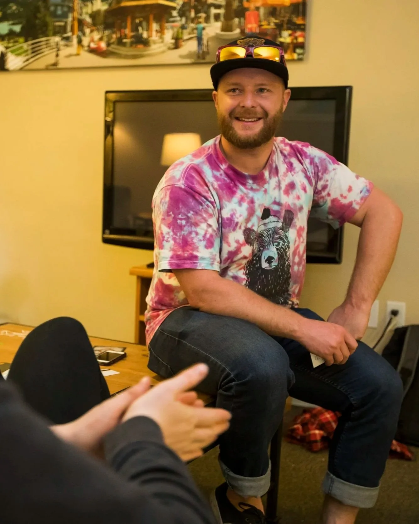 Sean - a man with a beard, wearing a tie-dye shirt with a bear graphic, a black cap with sunglasses on top - is sitting on a chair in a room with a TV and colorful artwork on the wall behind him, talking to and smiling at someone just off camera.