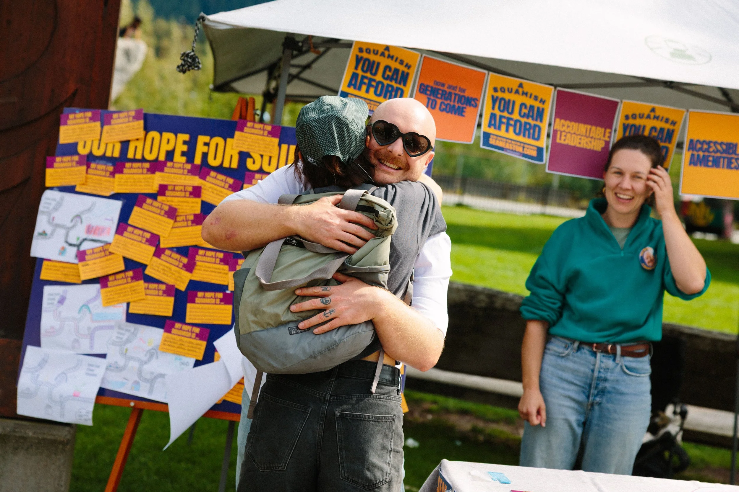 A photograph of Sean smiling and hugging someone who has their back to the camera. He is standing in front of a tent and poster with brightly coloured campaign material. Slogans say "A Squamish You Can Afford" and "Our Hope for Squamish"