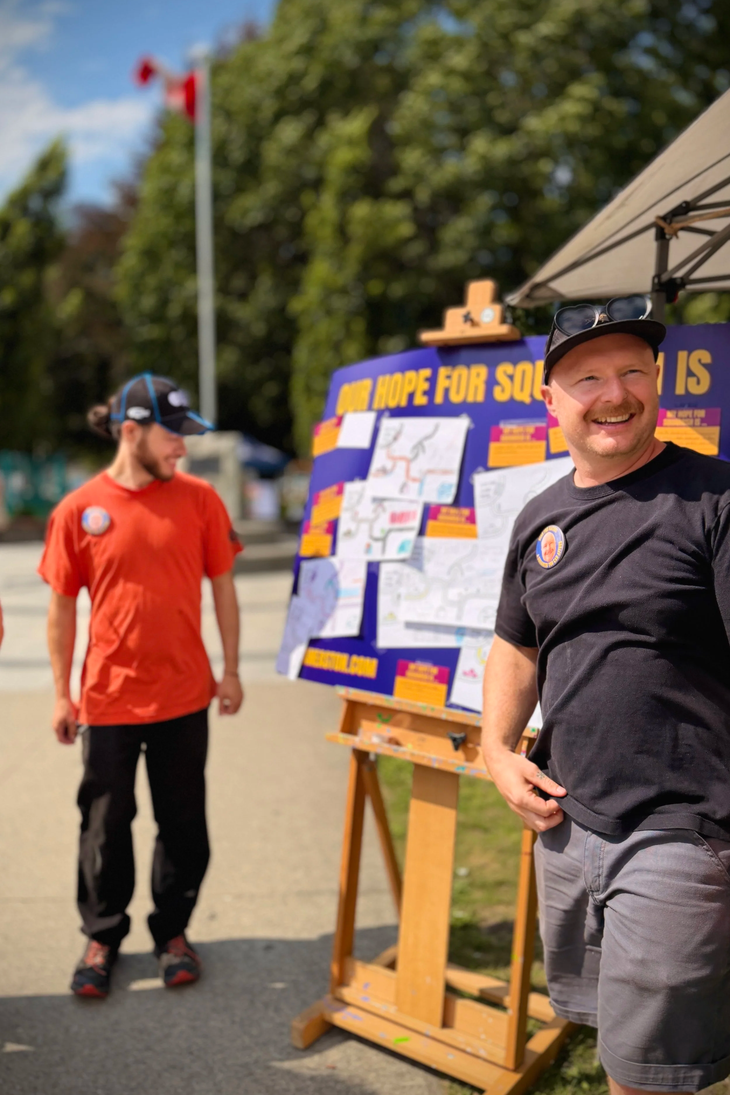 Two men are standing by a brightly coloured board that says "My hope for Squamish" with contributions from the community. In the foreground, Sean is laughing and looking off camera