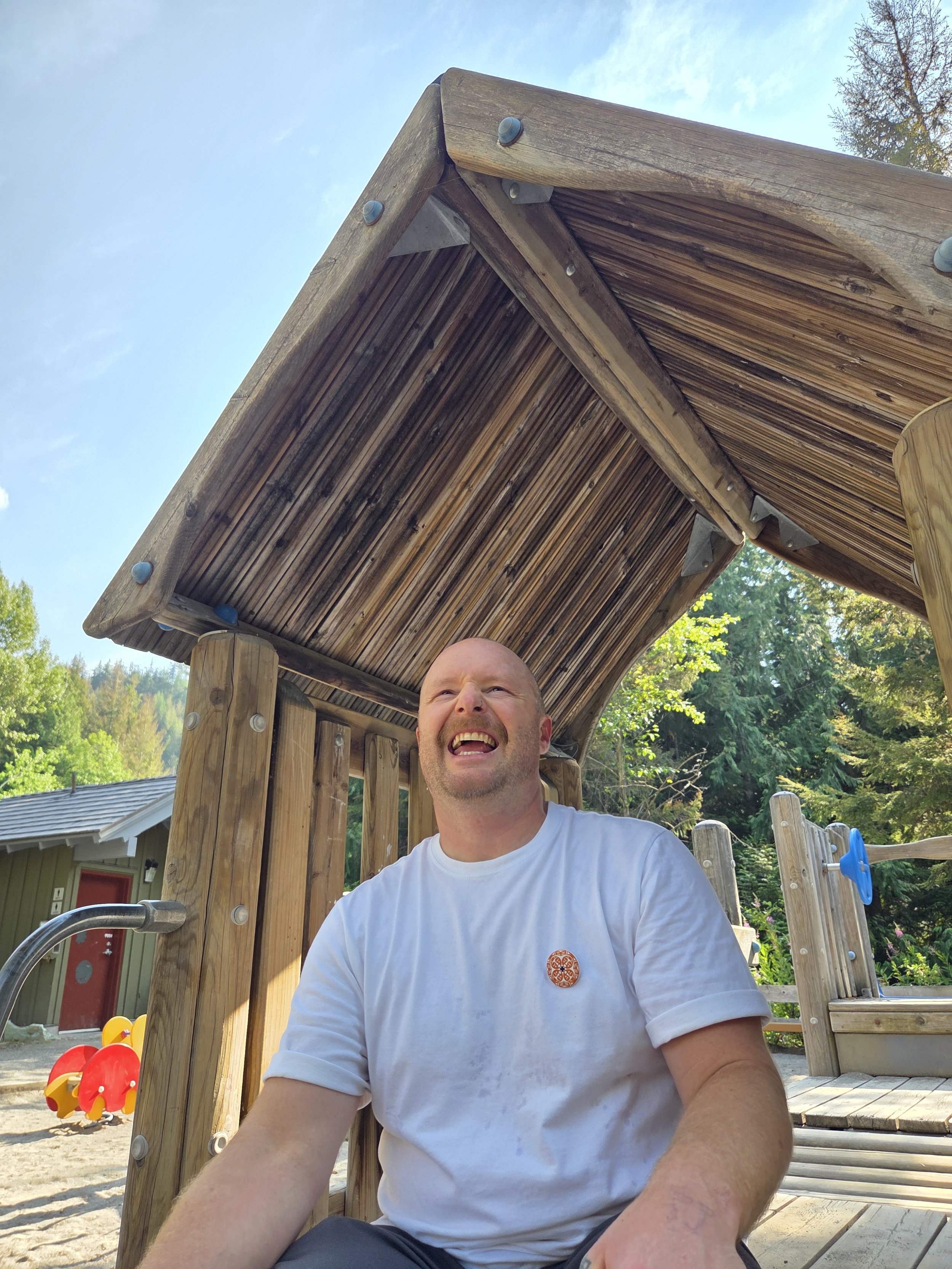 Sean, a man in his 30s, is wearing a white T-shirt, is sitting on a wooden play structure at a playground. The play structure is wooden and is outdoors surrounded by trees and a blue sky. Sean is smiling, with an expression of joy or surprise.