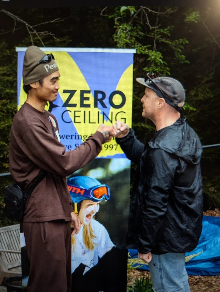Sean is fist bumping another young man in front of a banner that reads "Zero Ceiling". The youth is wearing a brown hoodie and beanie, and Sean is wearing a black jacket and cap. There are trees and outdoor scenery in the background.