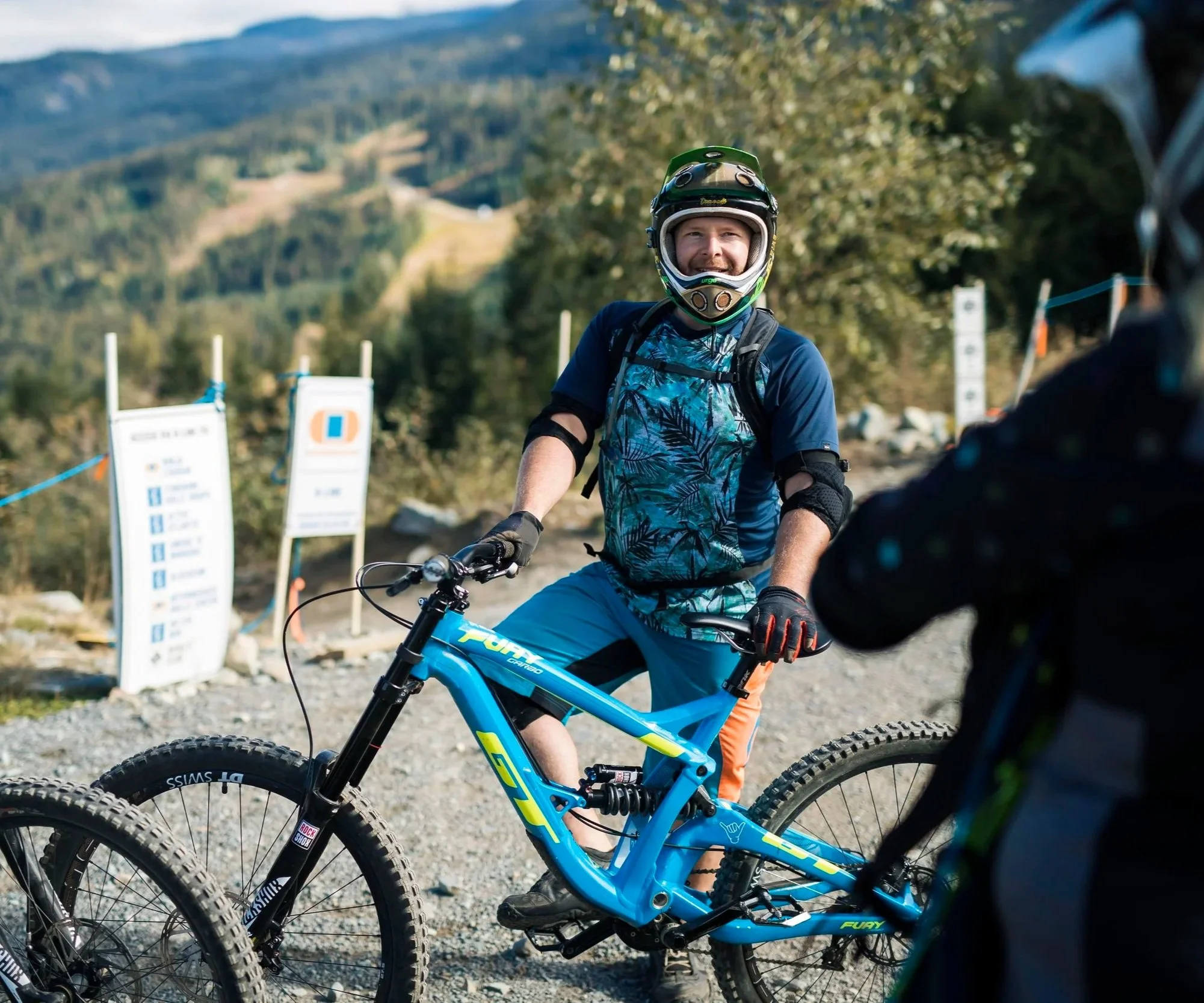 Sean is standing with a mountain bike outdoors with a scenic view of trees and hills in the background, wearing a helmet and a colorful shirt. He is talking with someone else who has their back to the camera, looking relaxed.