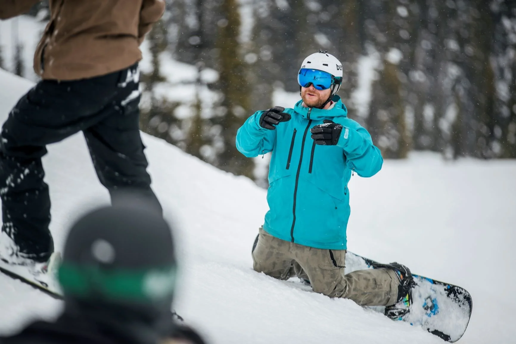 Sean is in a bright blue winter jacket, helmet, and goggles, kneeling on snow with a snowboard, talking to another person outdoors in a snowy forest. It appears he is providing coaching.