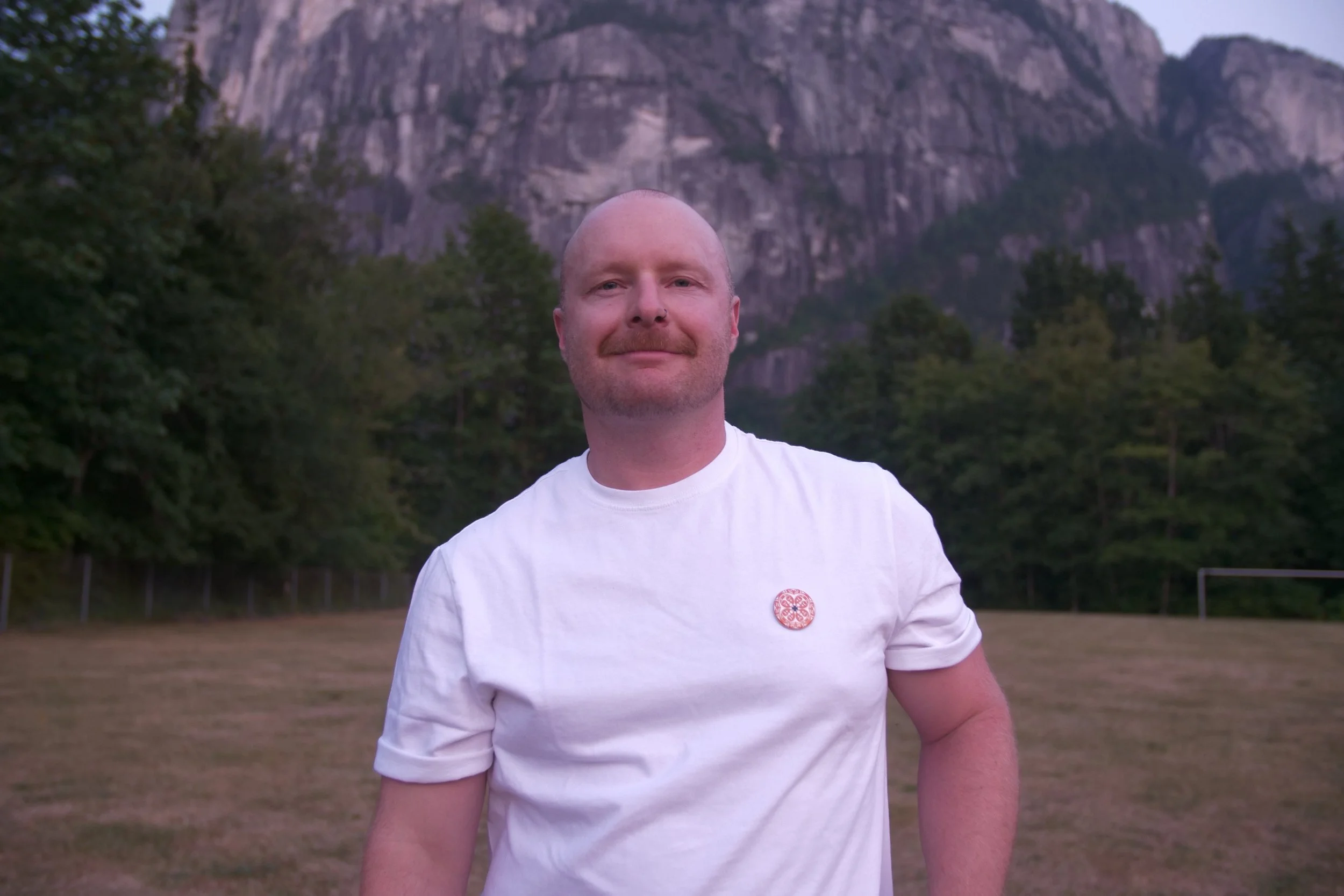 A photo Sean Easton. He is standing in front of the Squamish Chief mountain looking directly into the camera, wearing a white shirt and a pin.