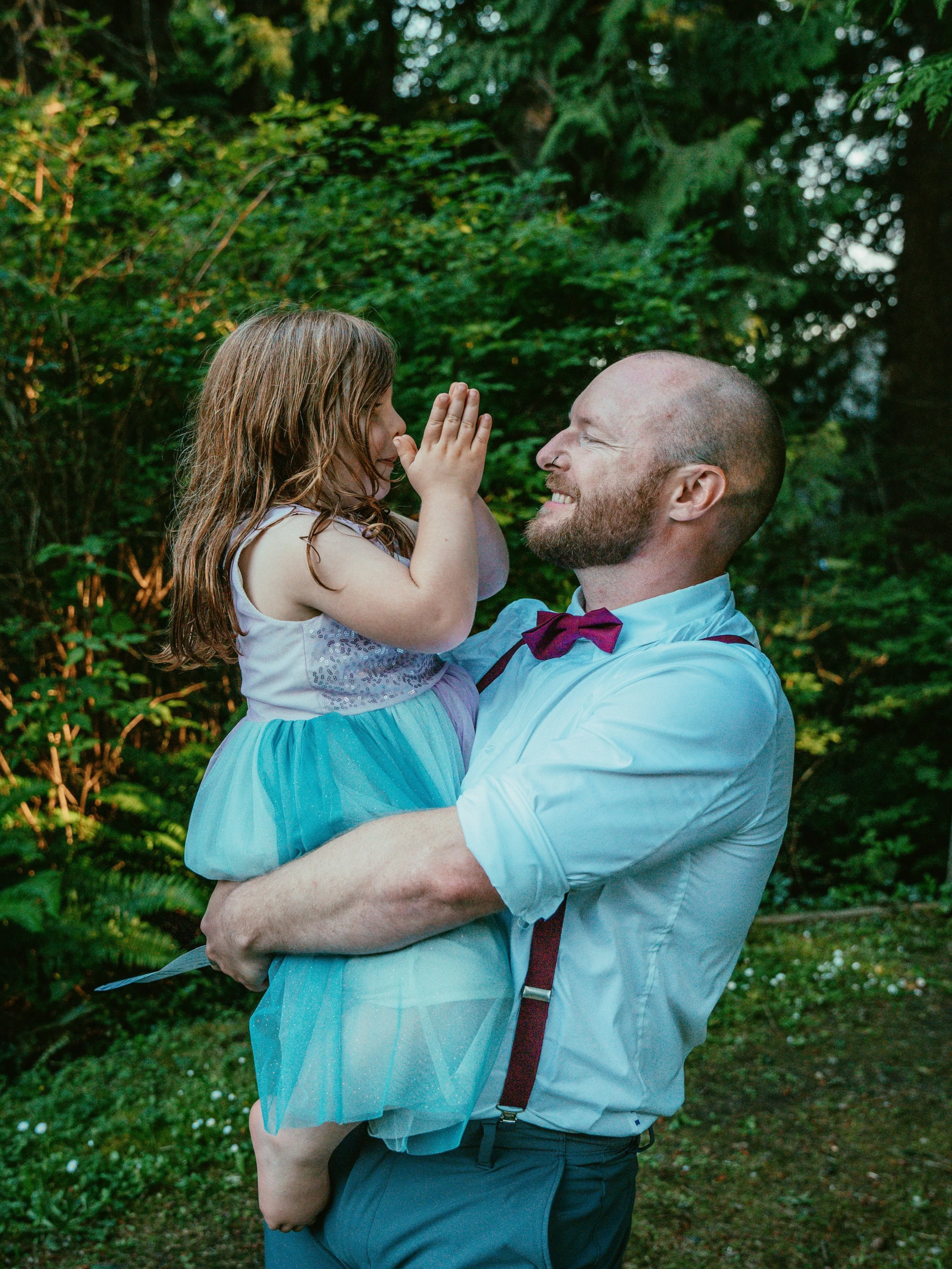 Sean, a man in his 30s, is holding his daughter, aged around 6,  in his arms. They are looking at each other, smiling, outdoors with lush green trees in the background. They are both dressed smart, as if they are guests at a wedding.